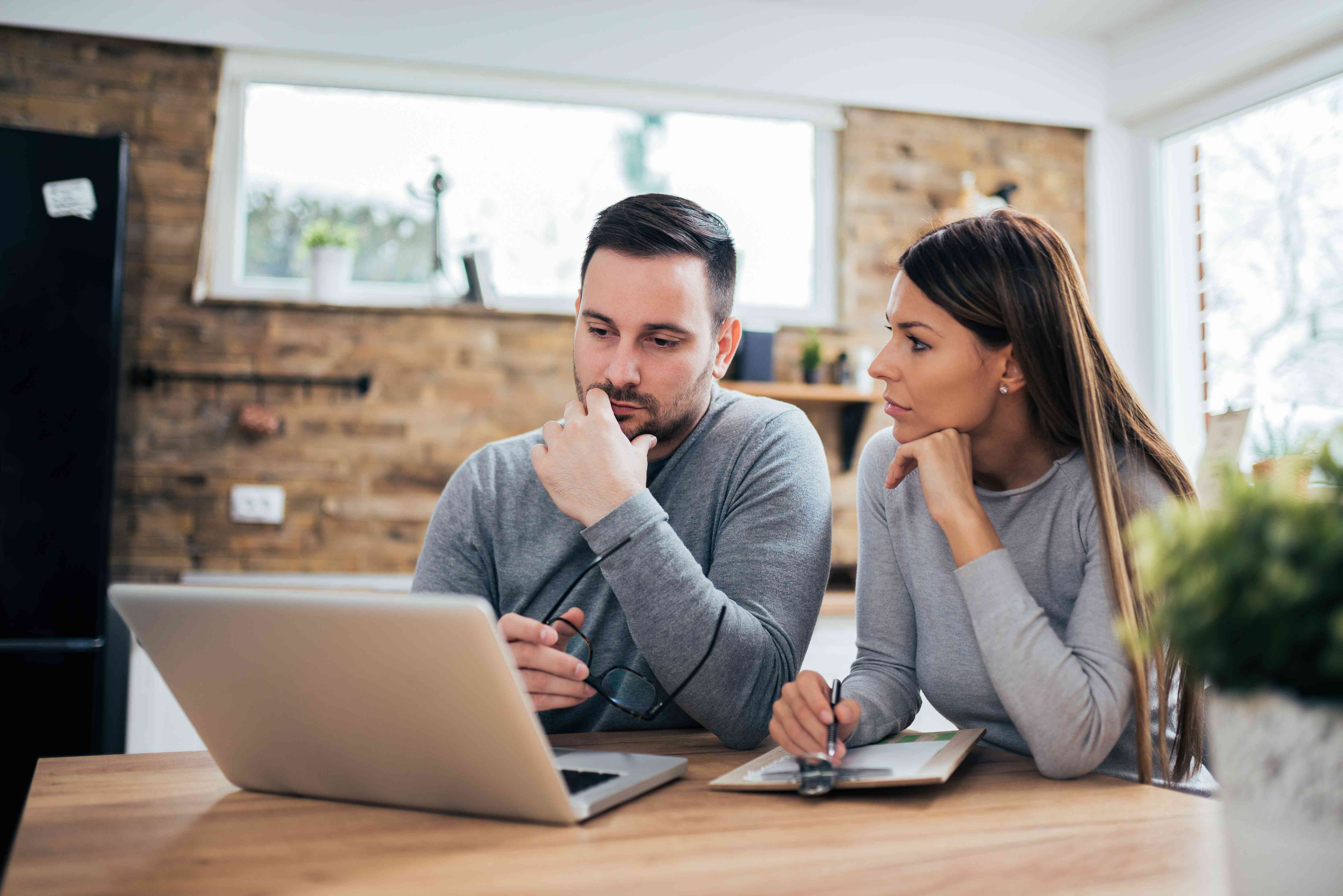 Couple in their 30s at their kitchen table looking together at a laptop