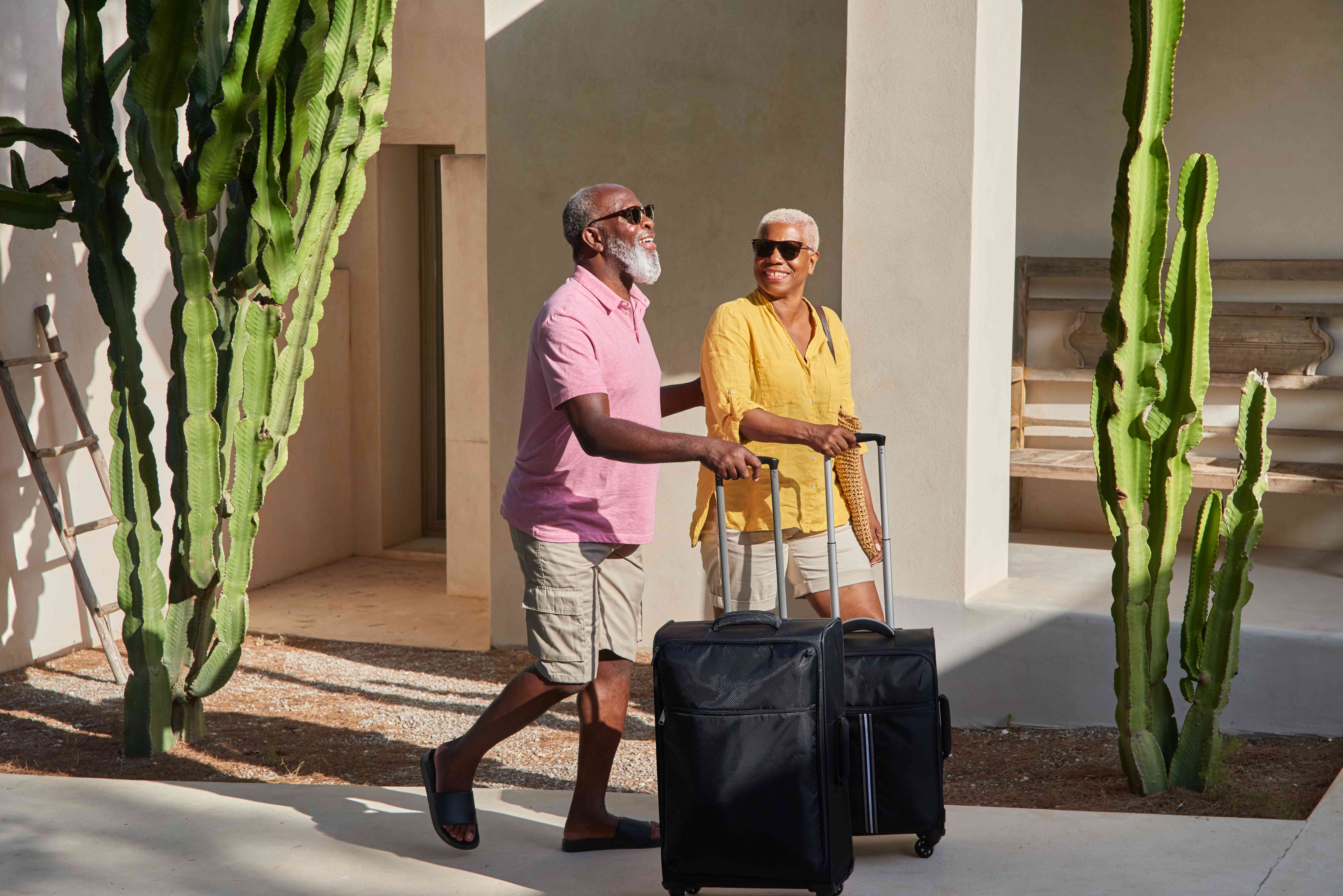 A man in a pink shirt and a woman in a yellow shirt, both wearing sunglasses, walk with luggage in a desert environment outside a house