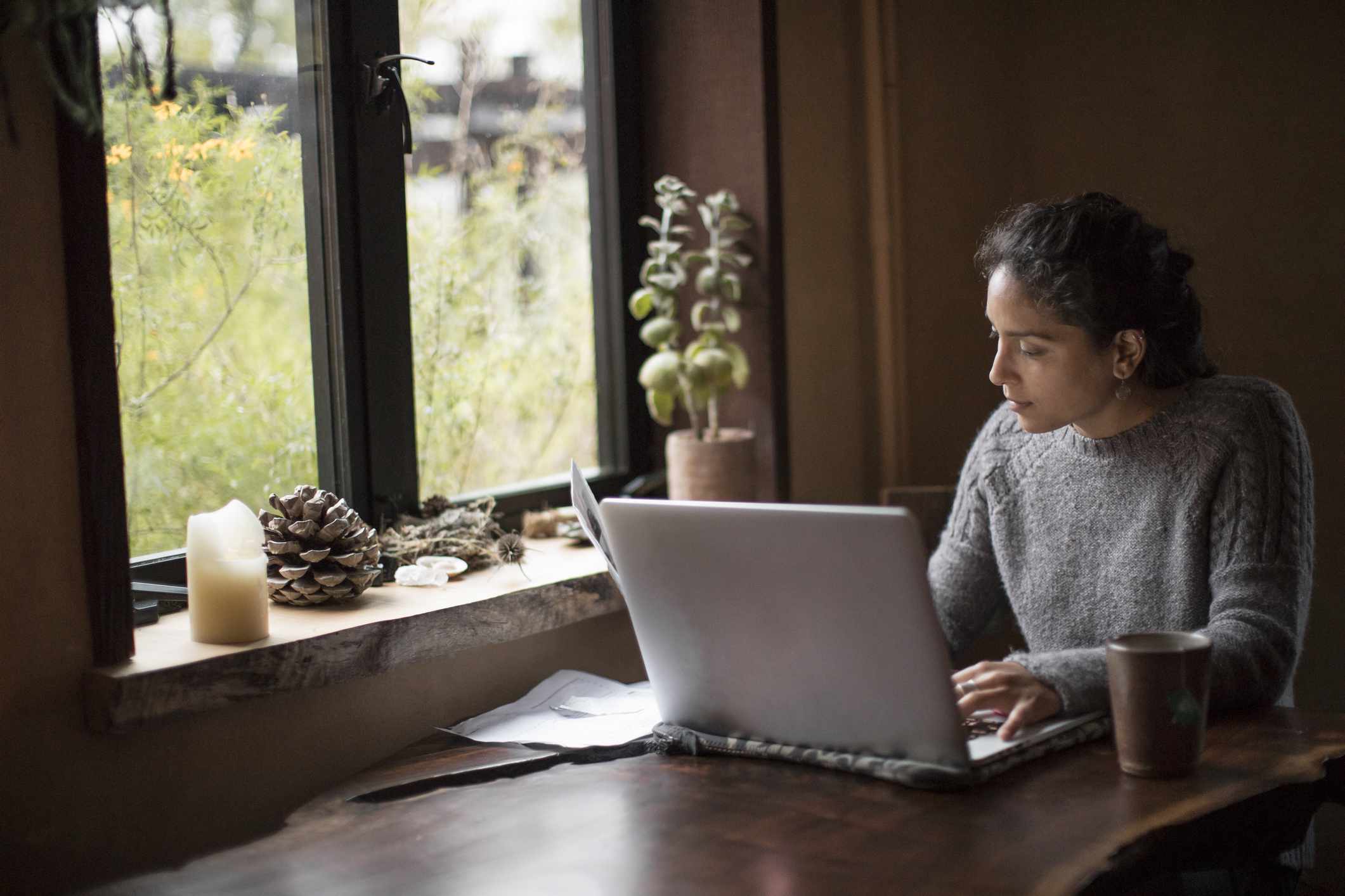 A woman sitting at a table by a window working on her laptop seriously