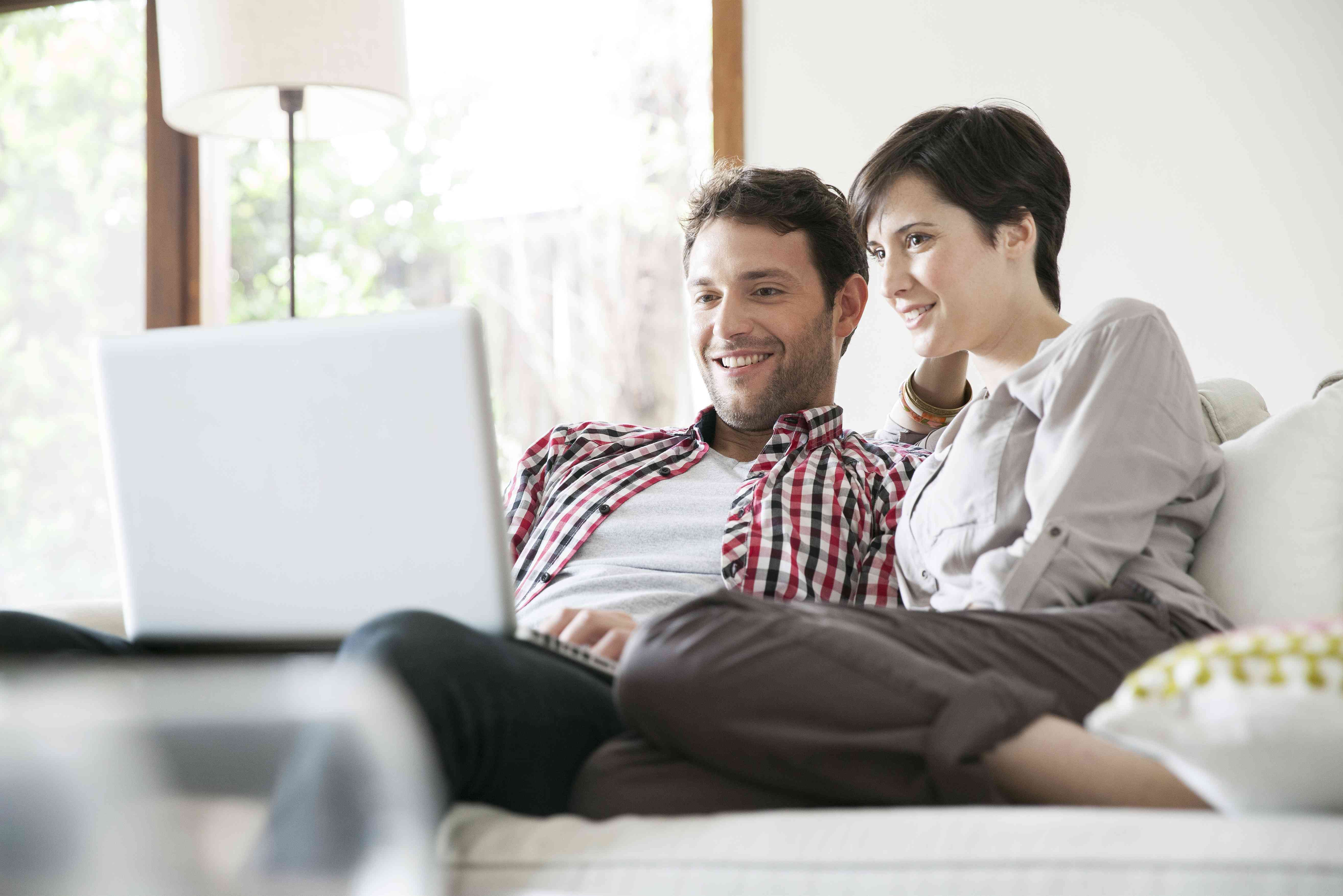 Couple in their 30s at home on the couch, looking together at a laptop and smiling