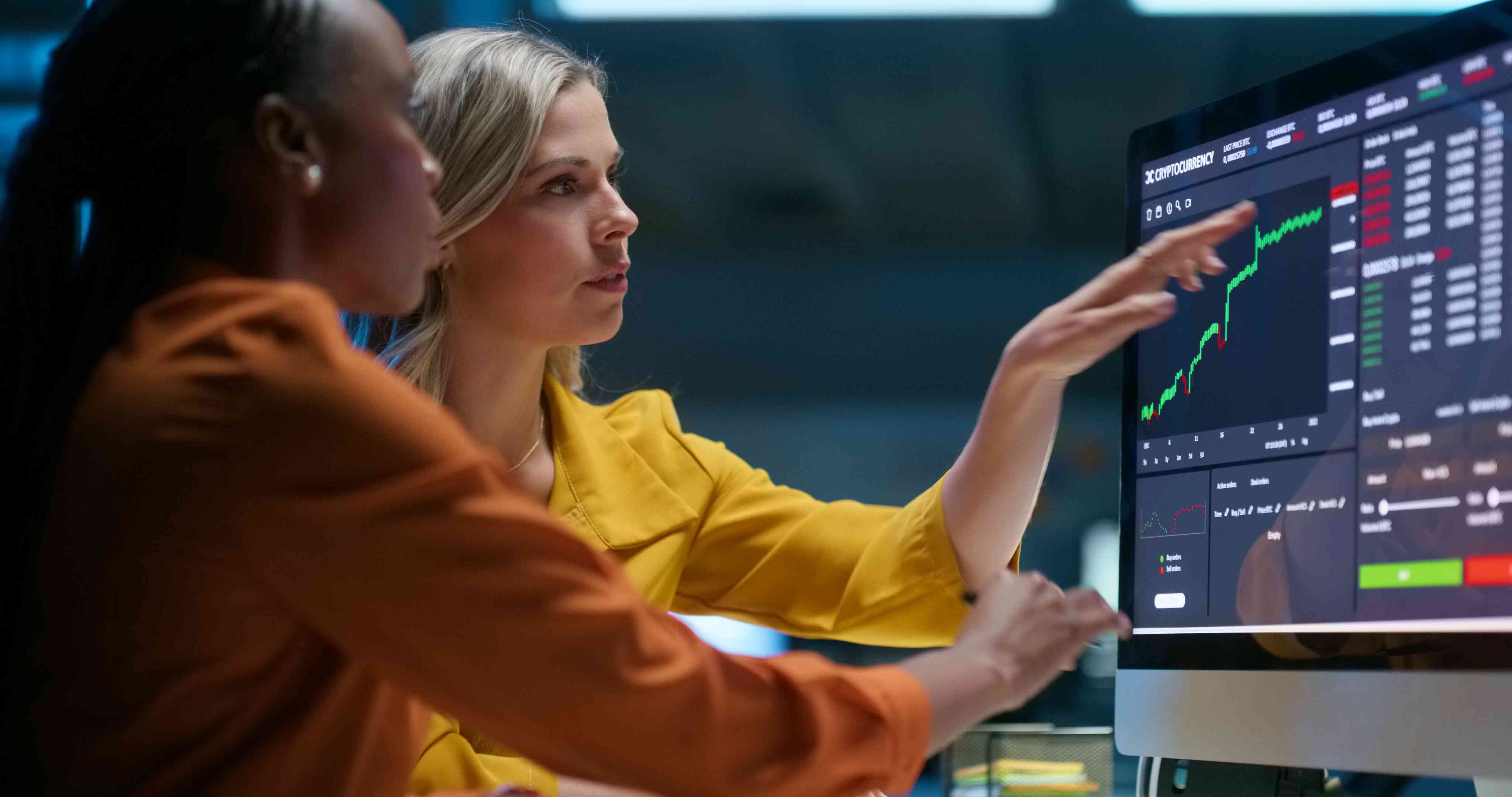 A businessperson points at a stock chart on a computer screen while another follows along.