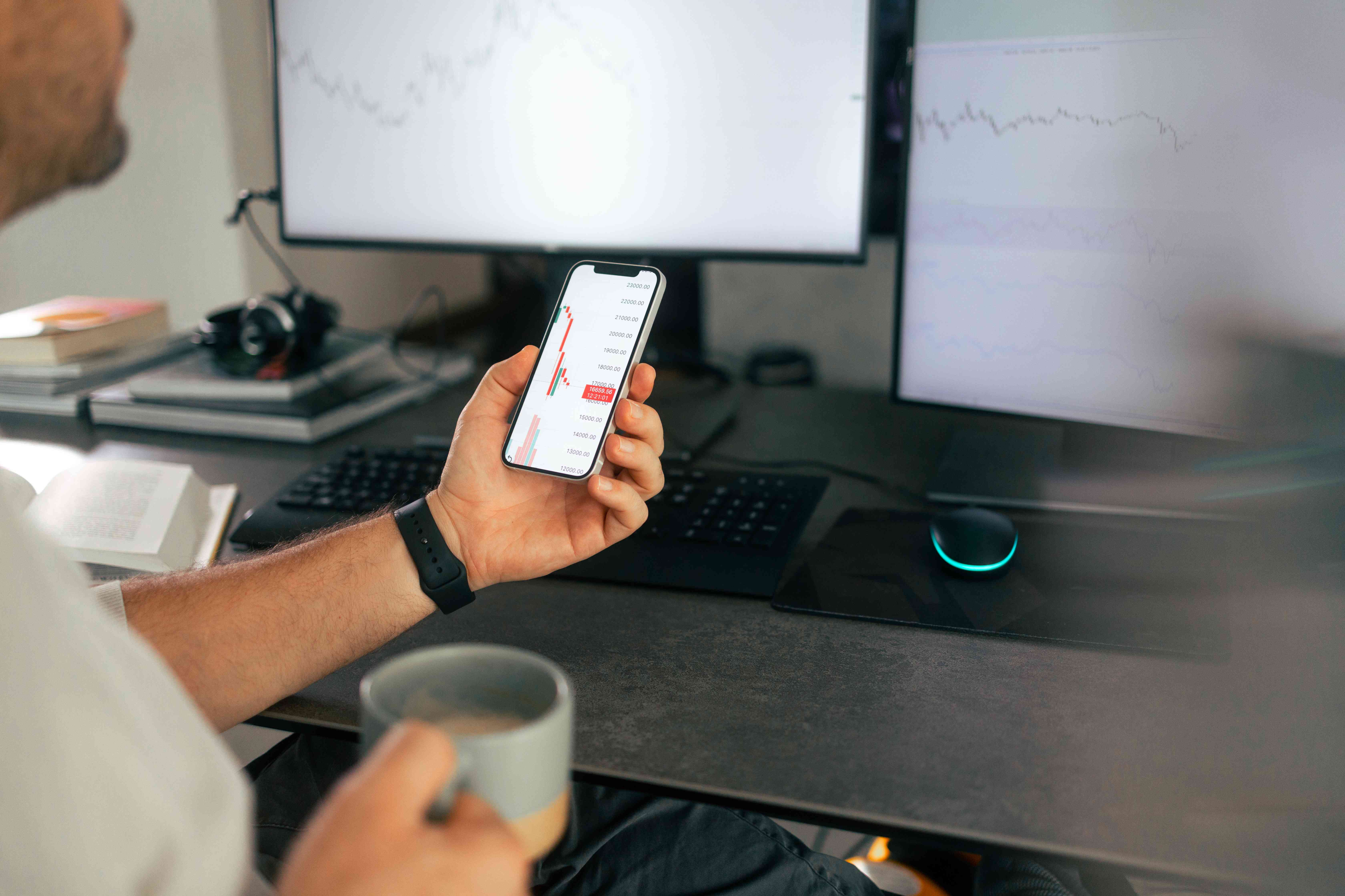 A man holds a phone with financial data in front of two computer screens