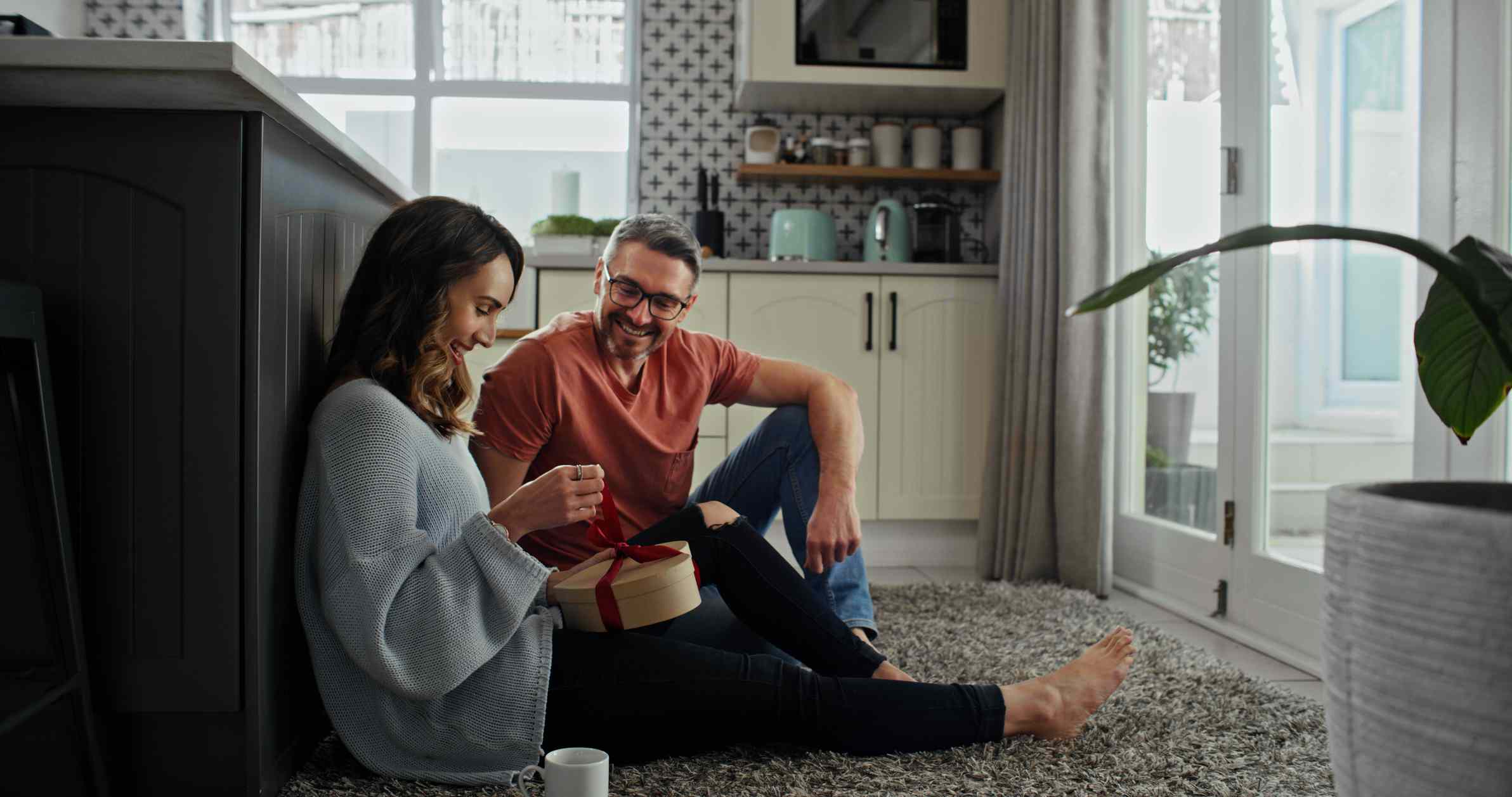 Woman opening a gift while man watches her, smiling, both sitting on the kitchen floor