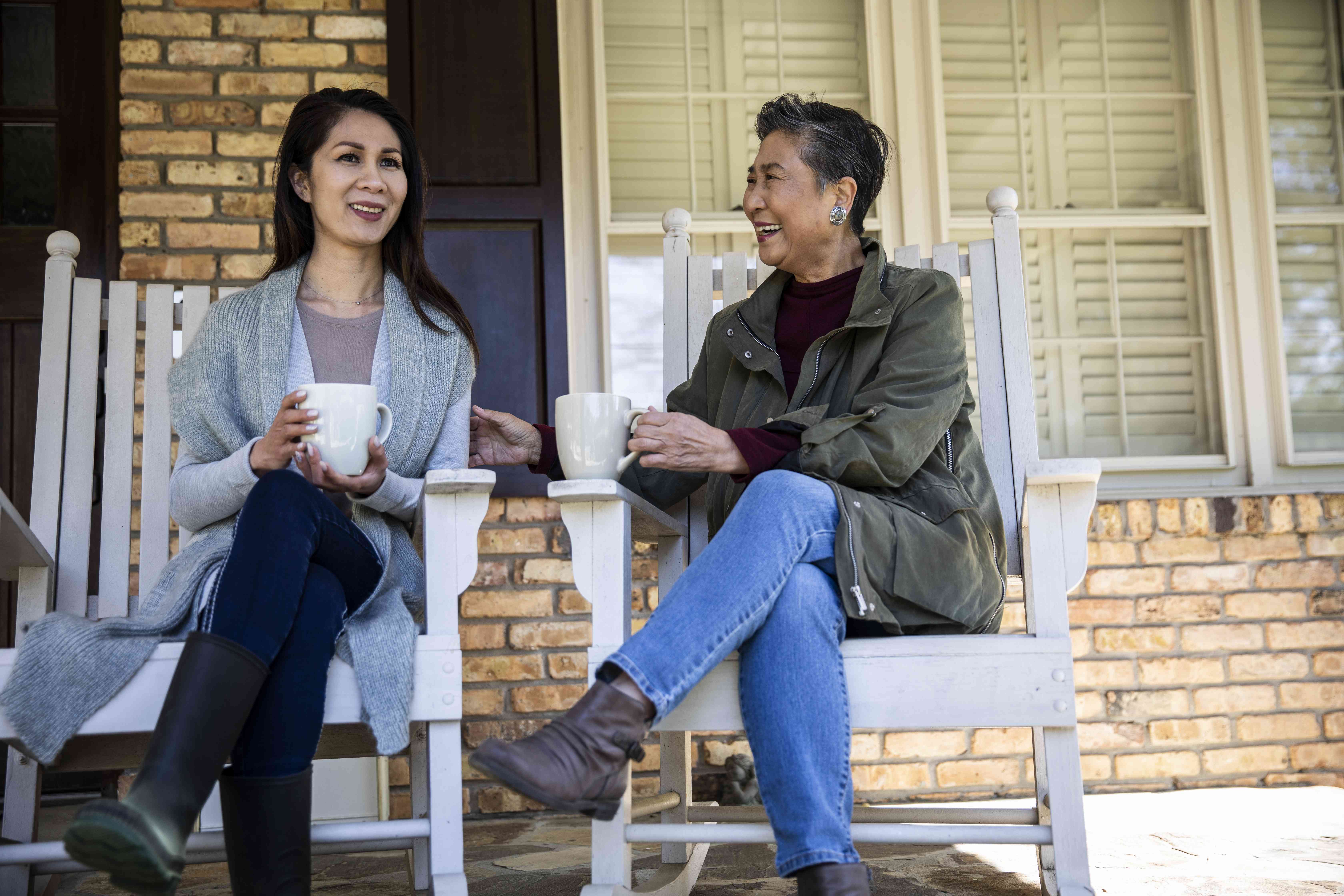 Two woman sit in chairs on a front porch, drinking out of mugs and talking