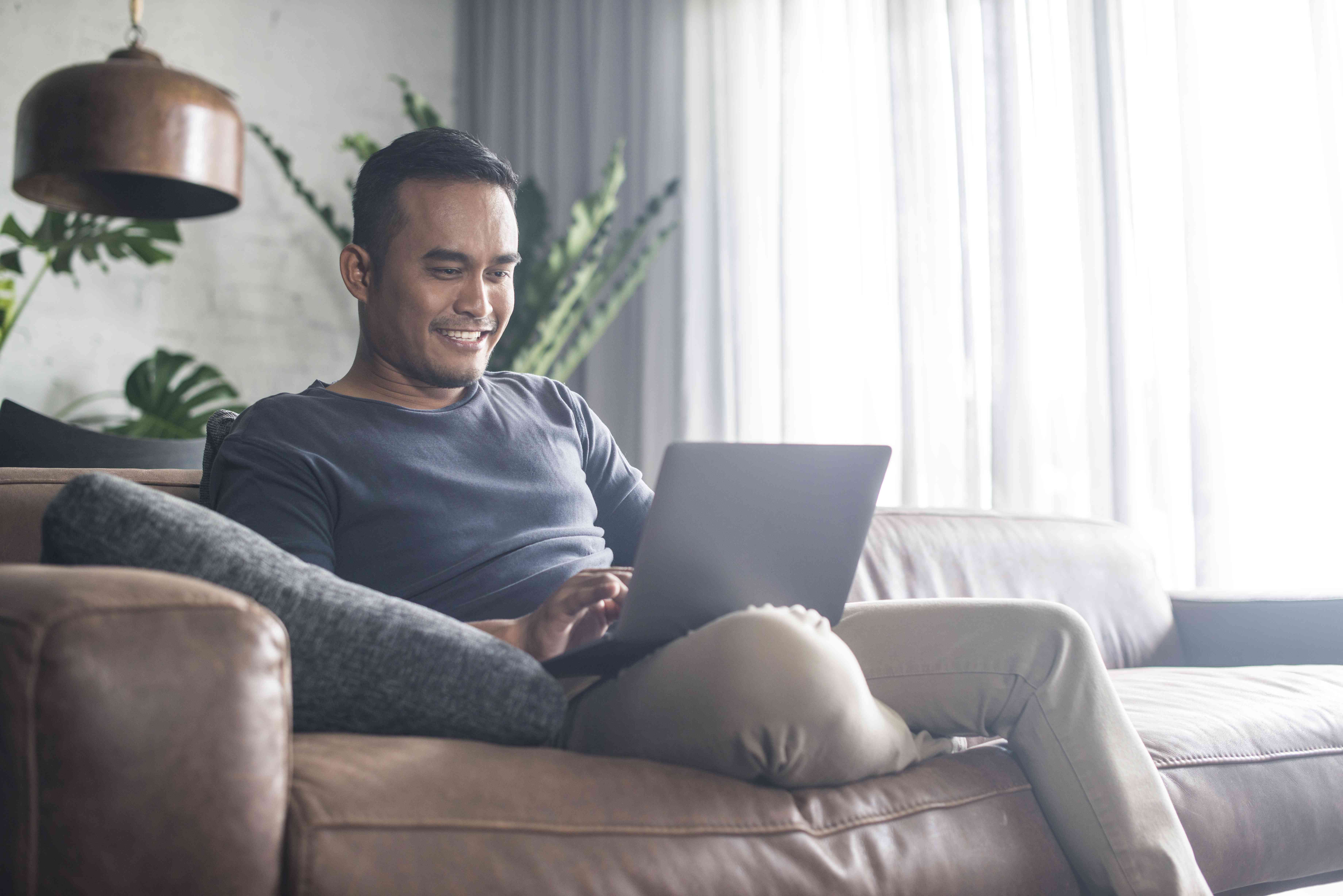 Man in his 30s sitting on his couch and smiling as he looks at his laptop