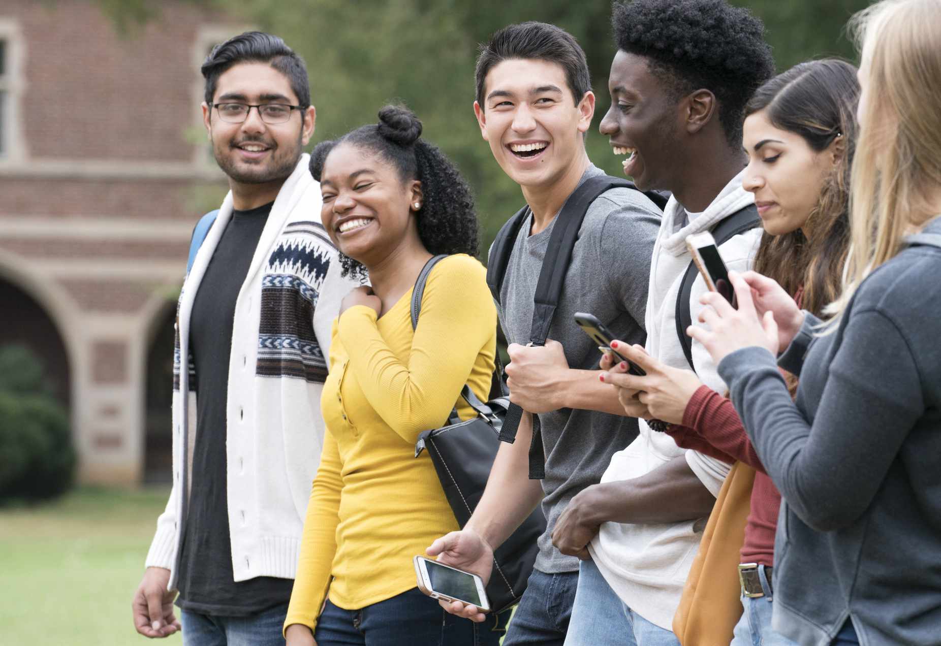 Six college students walk on campus. Four are laughing, one looks at her phone, and one has her back to the camera.