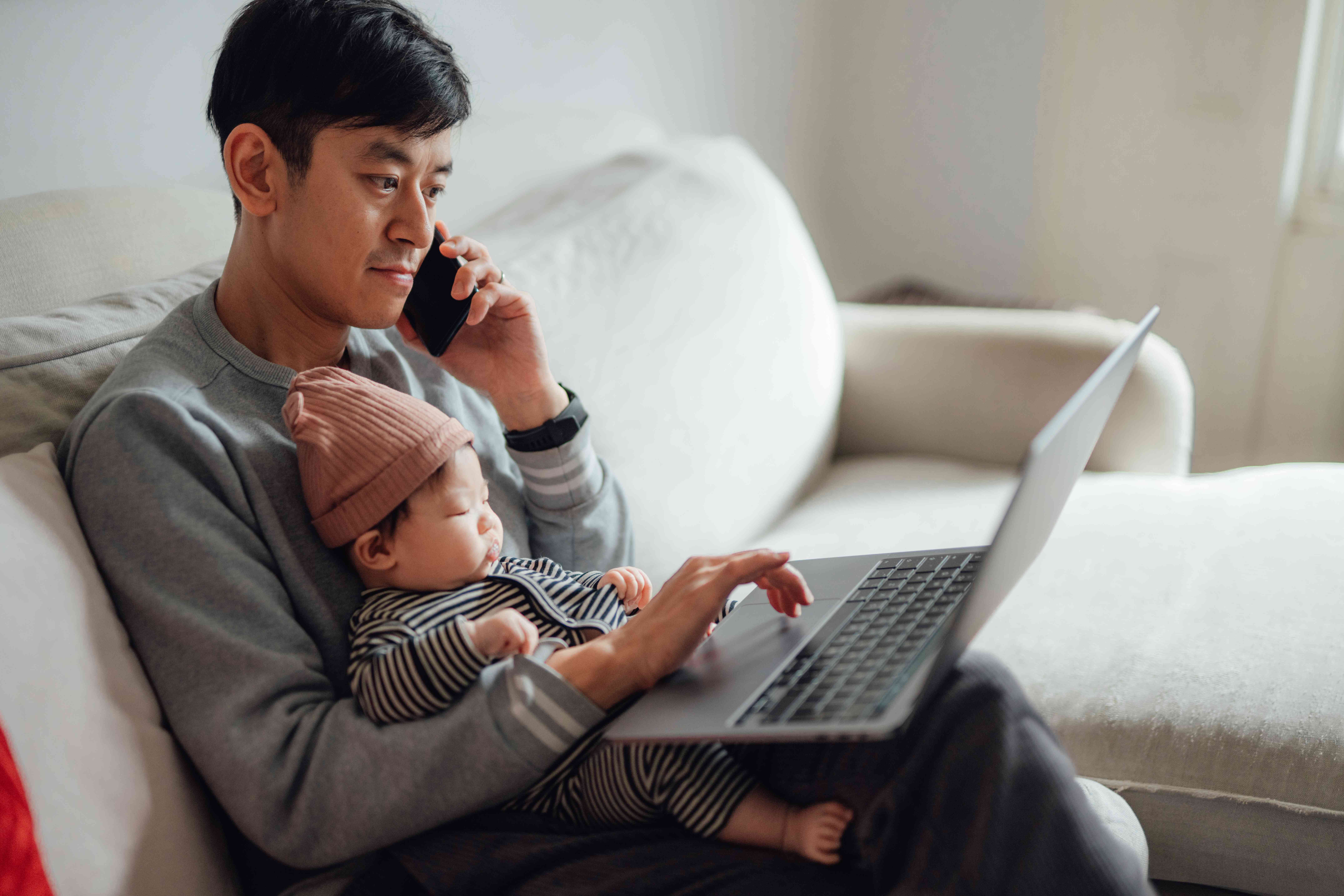 Asian man in his 30s sitting on his couch with a baby as he looks at a laptop and holds a phone to his ear