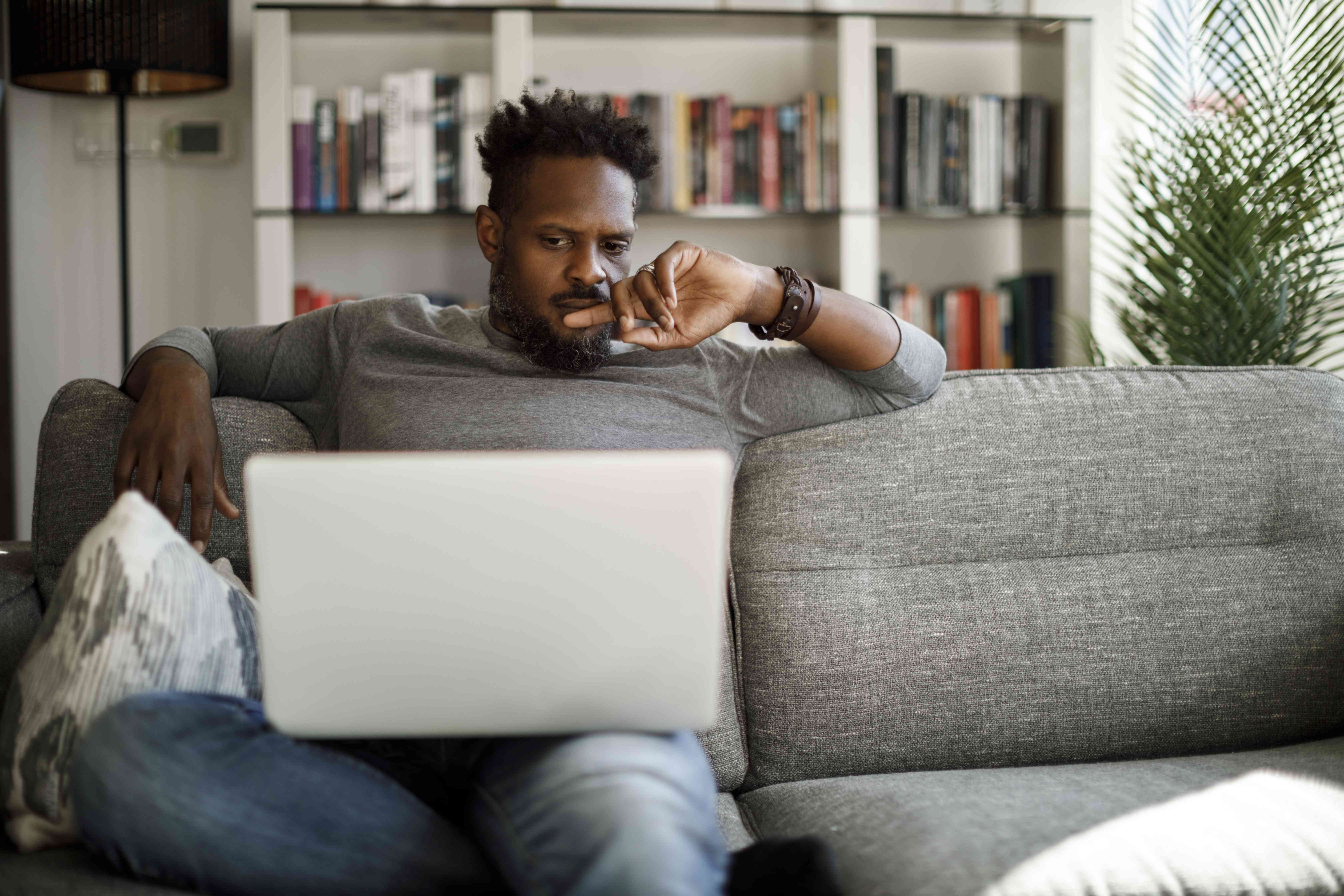 Man in his 30s on his living room couch seriously contemplating his laptop screen