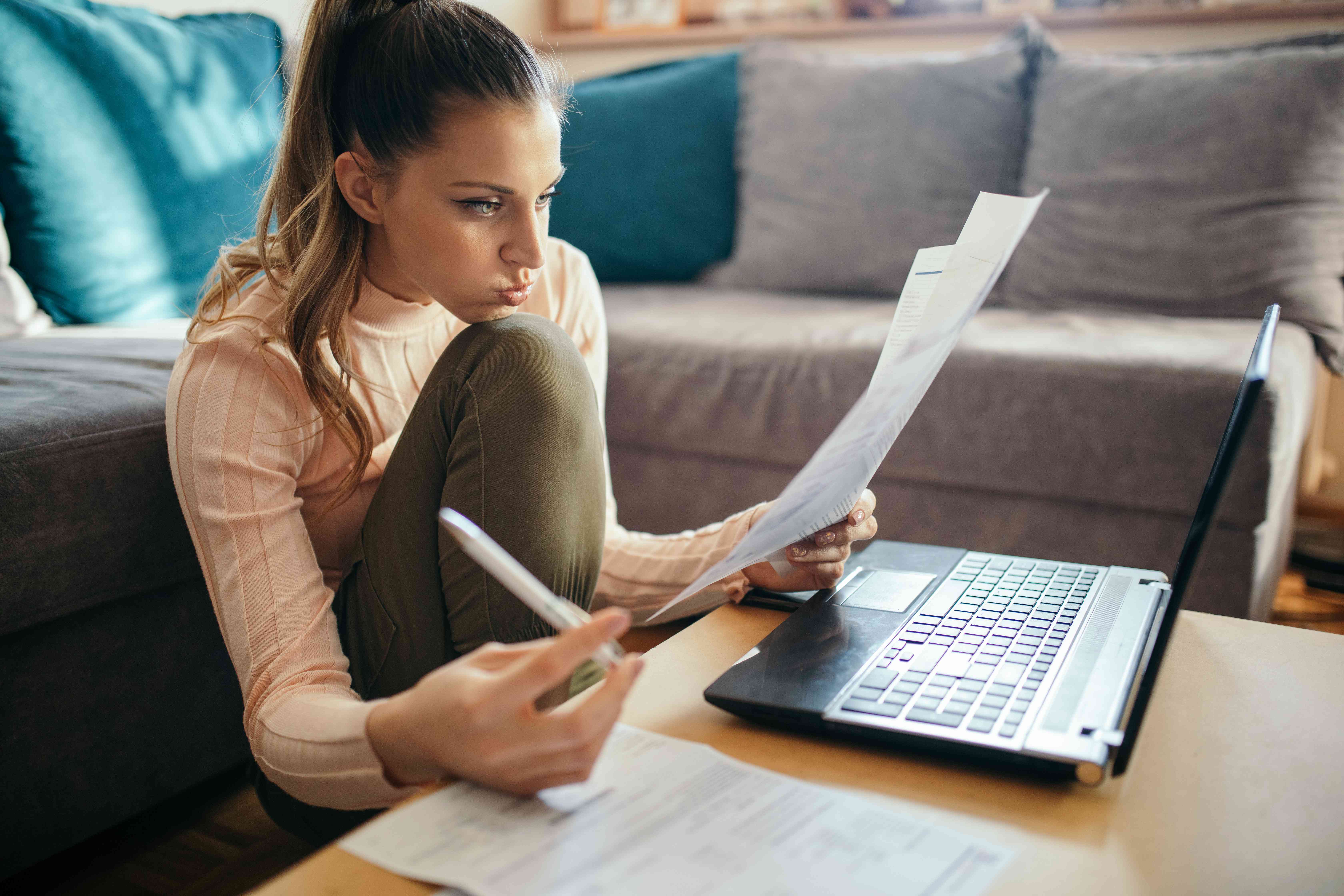 Young woman sitting on the floor next to her sofa with her laptop and bills, calculating her expenses