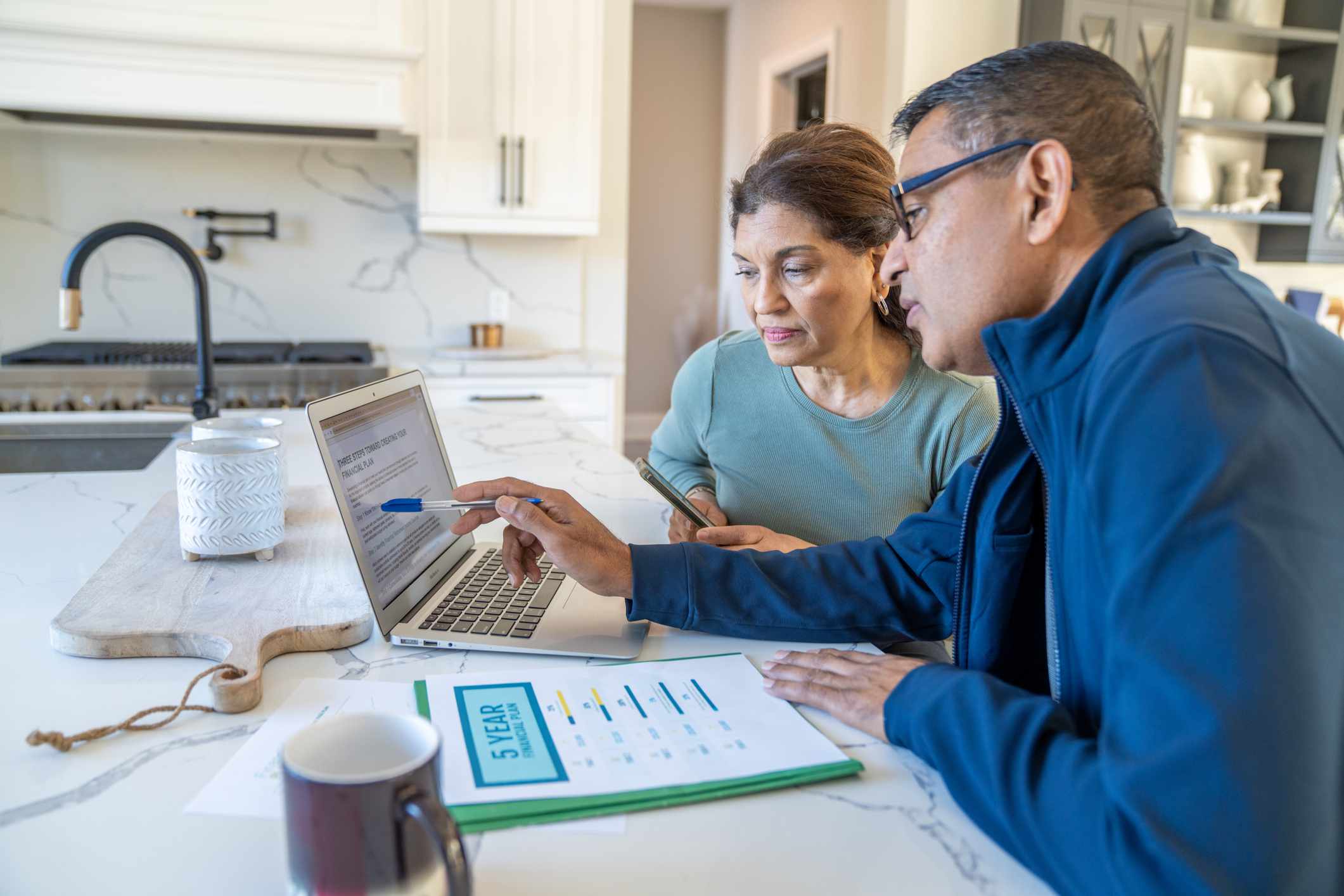 A couple sit at their kitchen island with a laptop open and their bills spread out in front of them as they review their finances together.