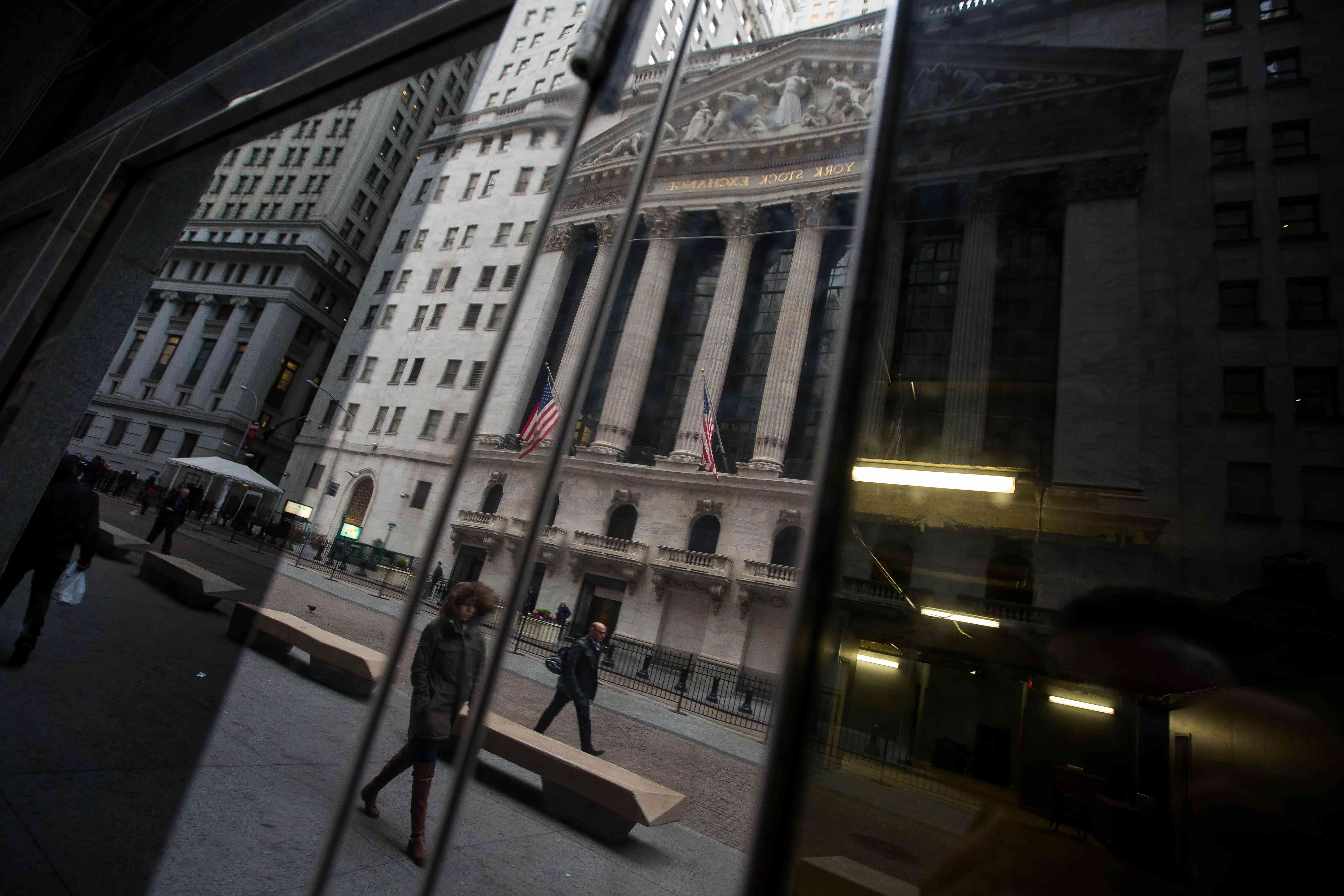 The New York Stock Exchange is reflected in a mirrored doorway, as people walk by. 