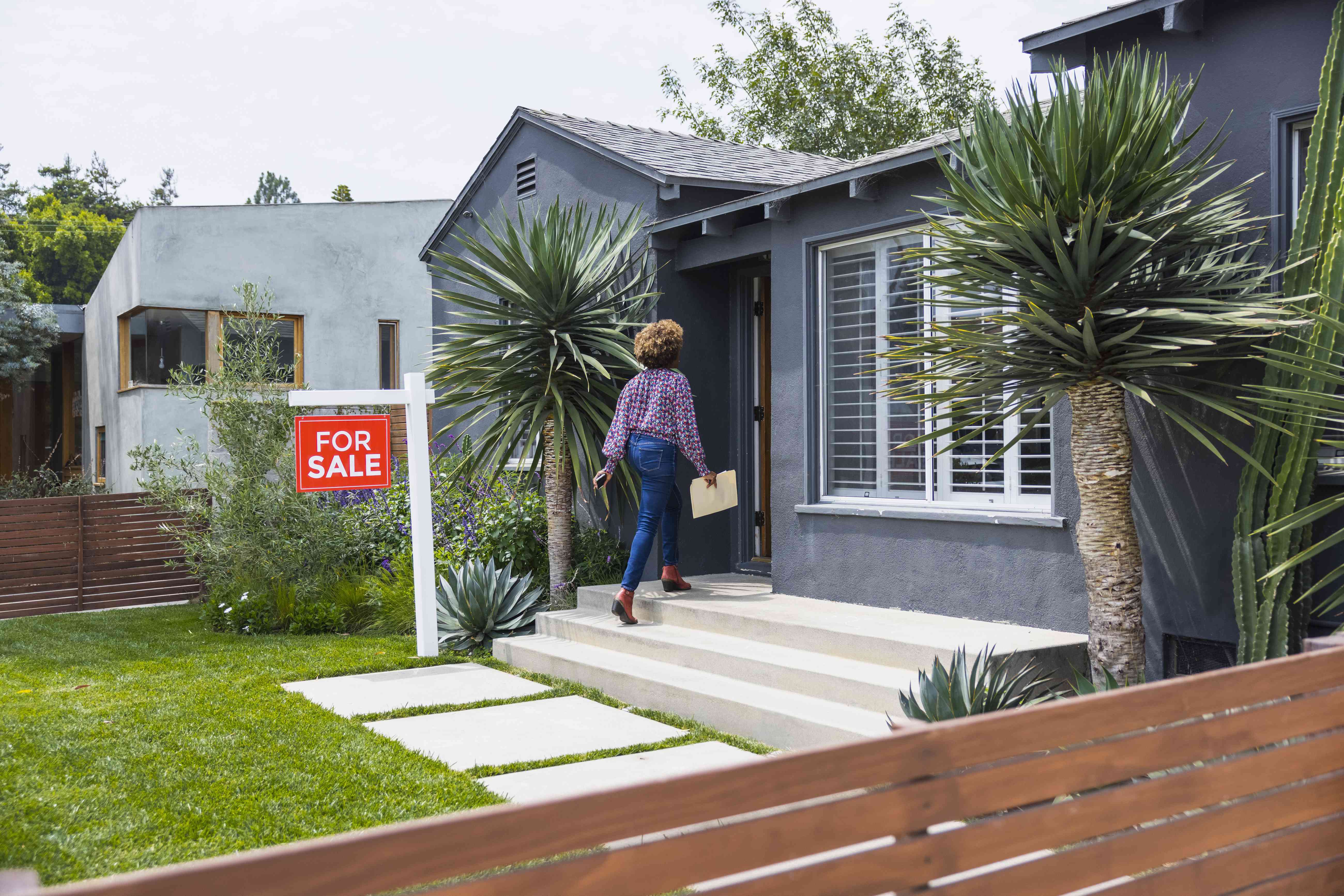 Woman walking into a home that has a "for sale" sign out front.