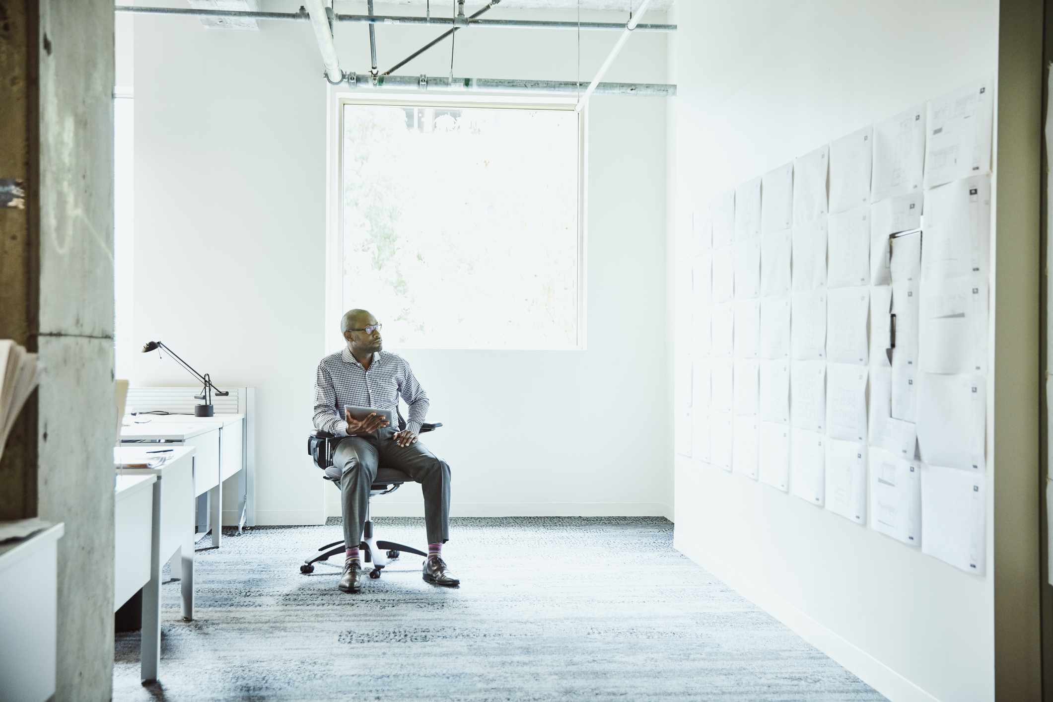 businessman sitting on a chair in an office looking at a wall full of papers while holding a tablet