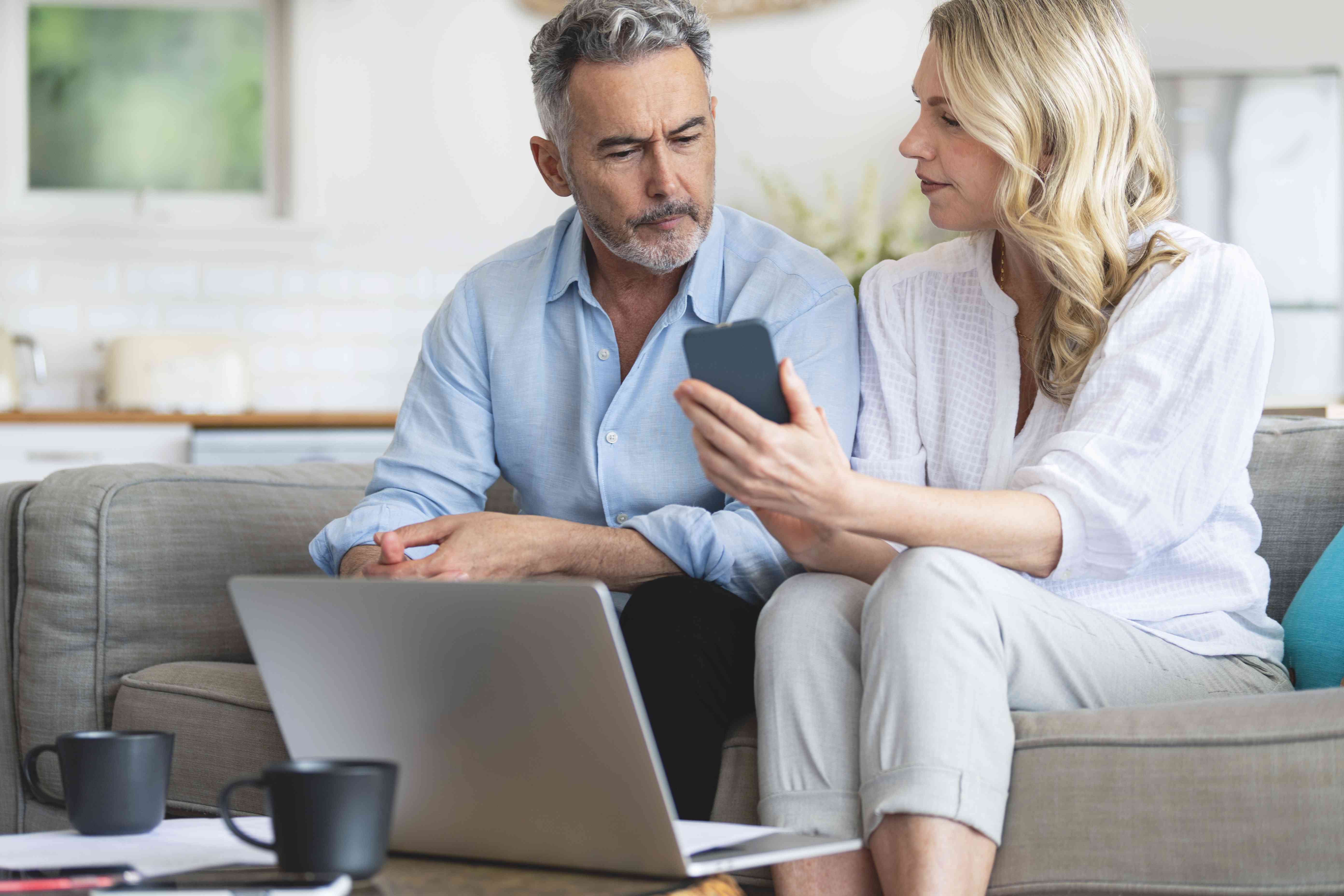 Older couple sitting on couch at home, looking together at a laptop and her smartphone.