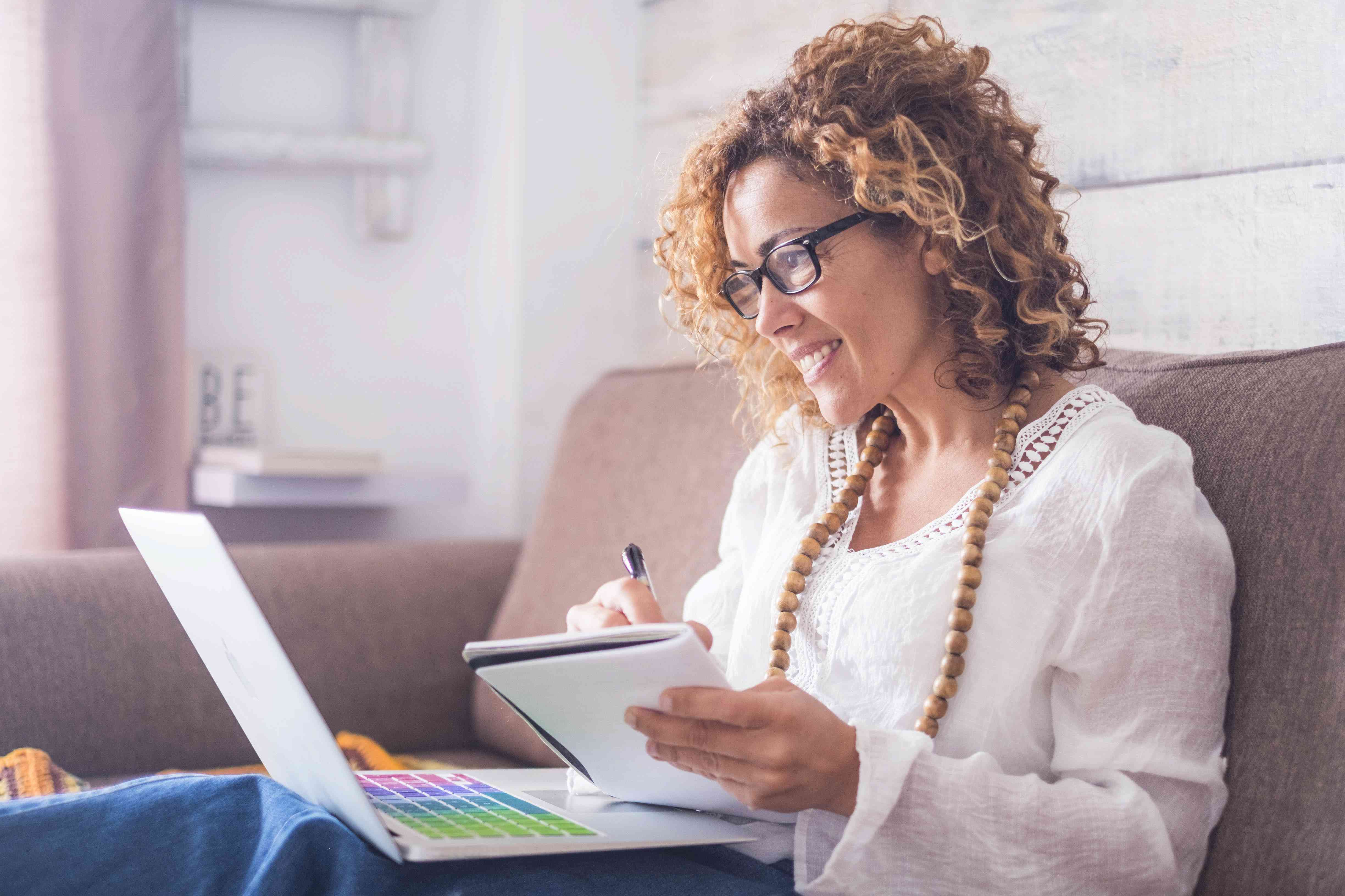 Woman in her 40s on her living room couch looking at her laptop with a pen and notebook in hand