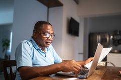 Older Black man at a kitchen table with a laptop holding paperwork