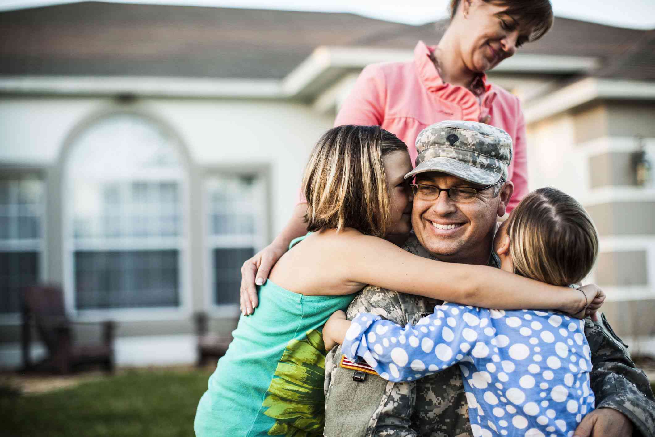 Mother watching over two daughters hugging their dad in military fatigues uniform in the front yard of their house.