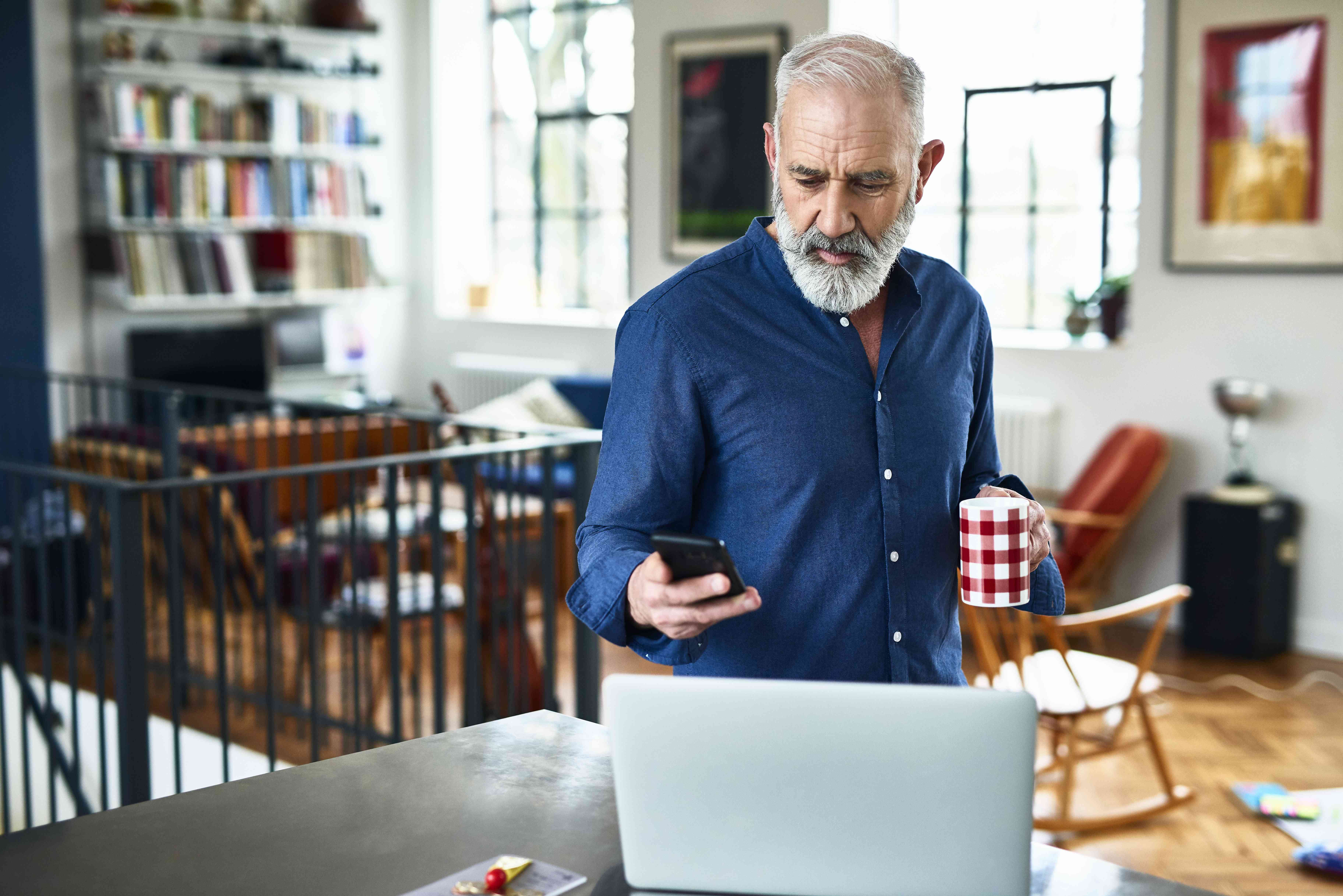 Older man at home, looking at his smartphone with a laptop open on kitchen counter