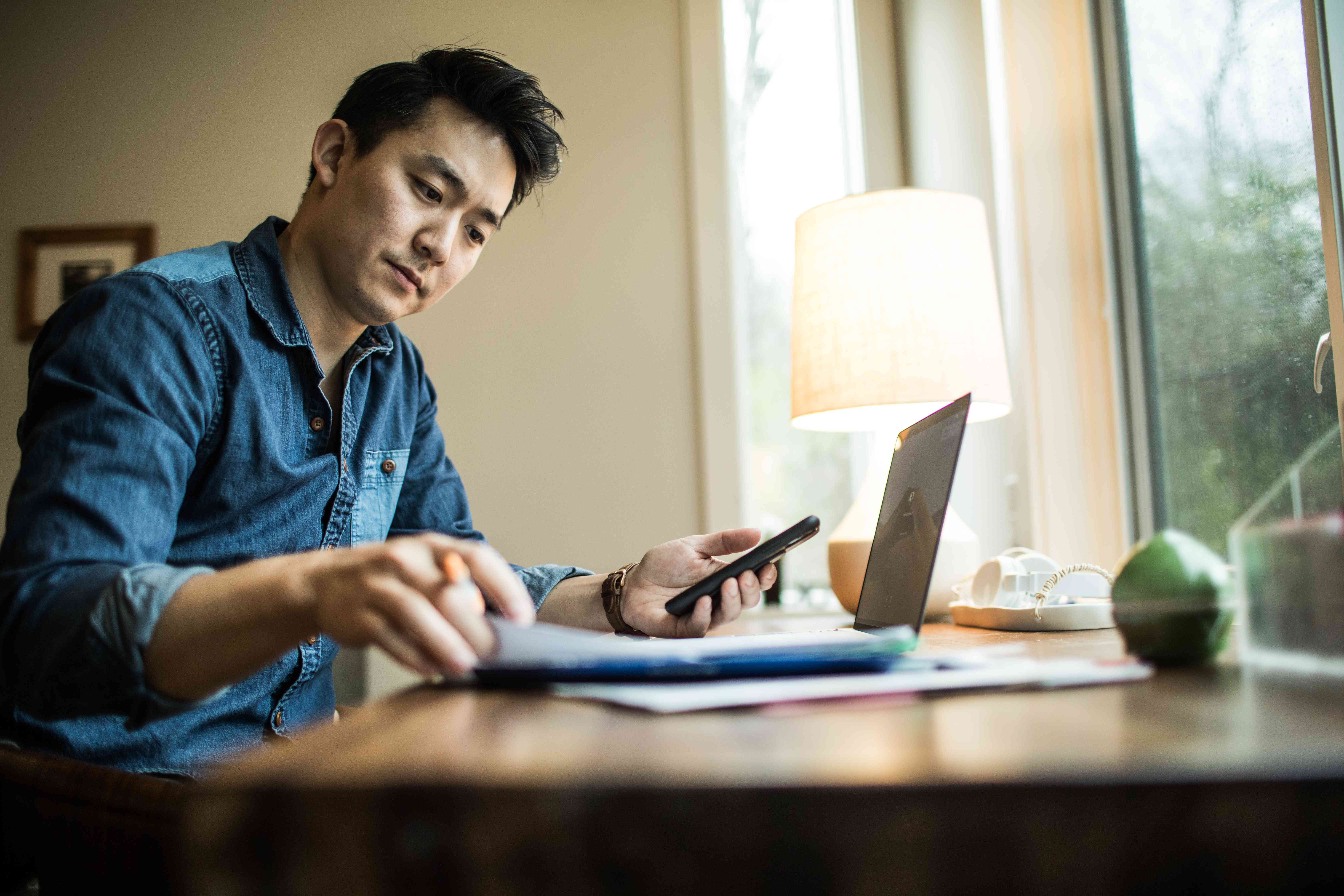 Man in his 30s at his desk at home, looking at financial documents while sitting in front of a laptop and holding his smartphone