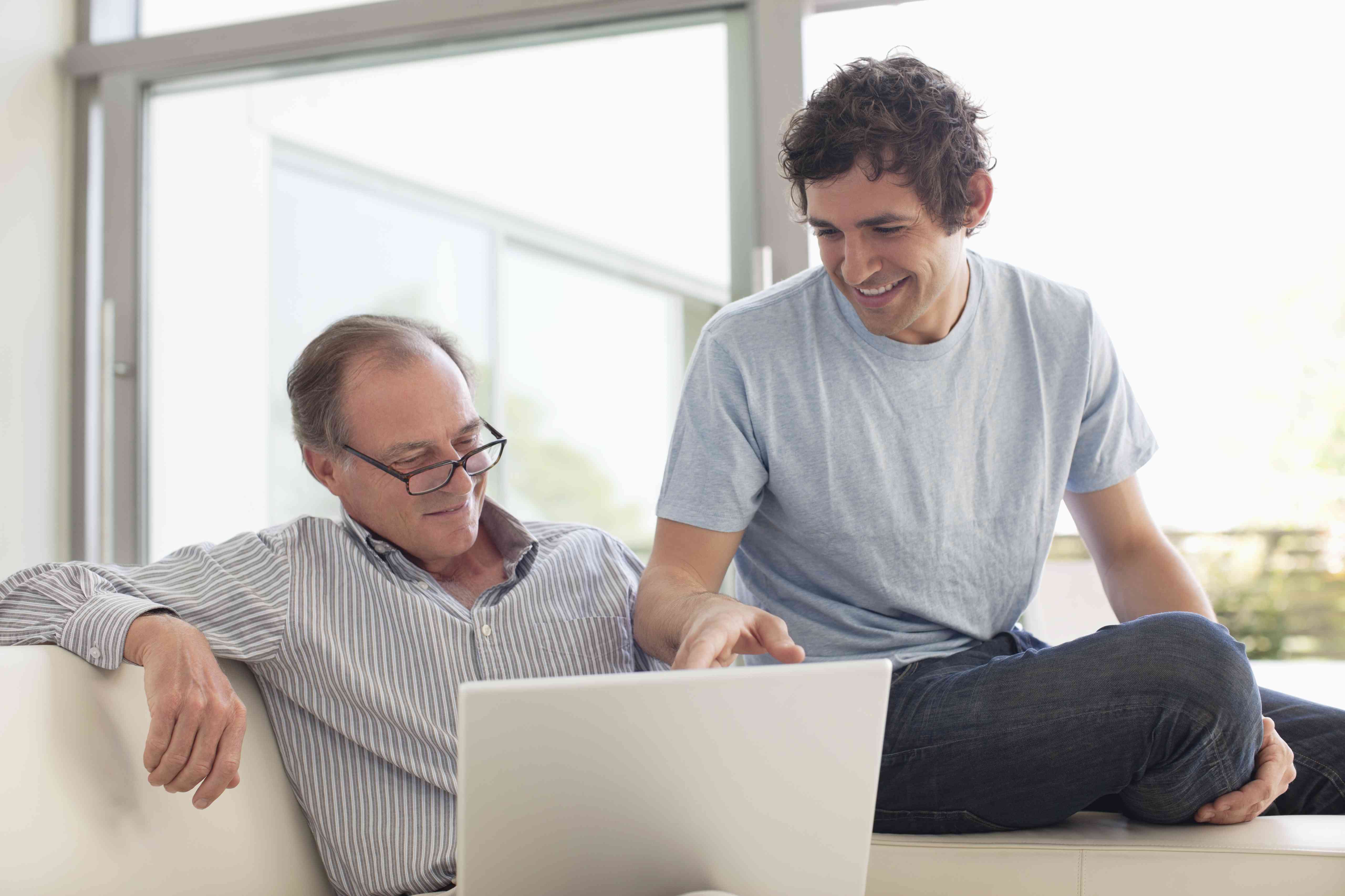 An older man and a younger man sit on a couch together looking at a laptop