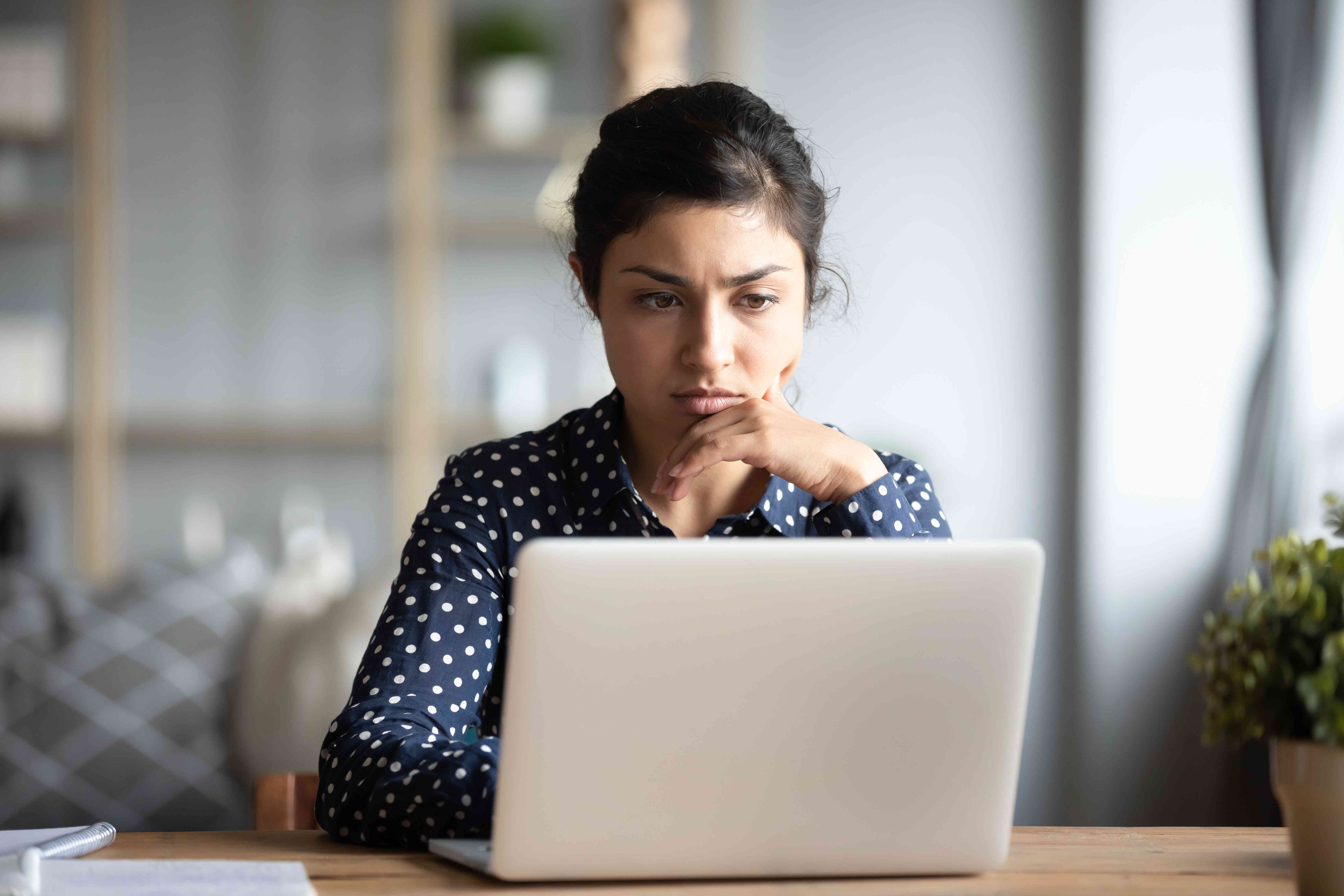 Young woman staring intently at her laptop screen while sitting at her dining room table