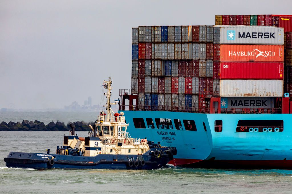 A cargo ship in the water carrying containers with a smaller tug boat on the side.