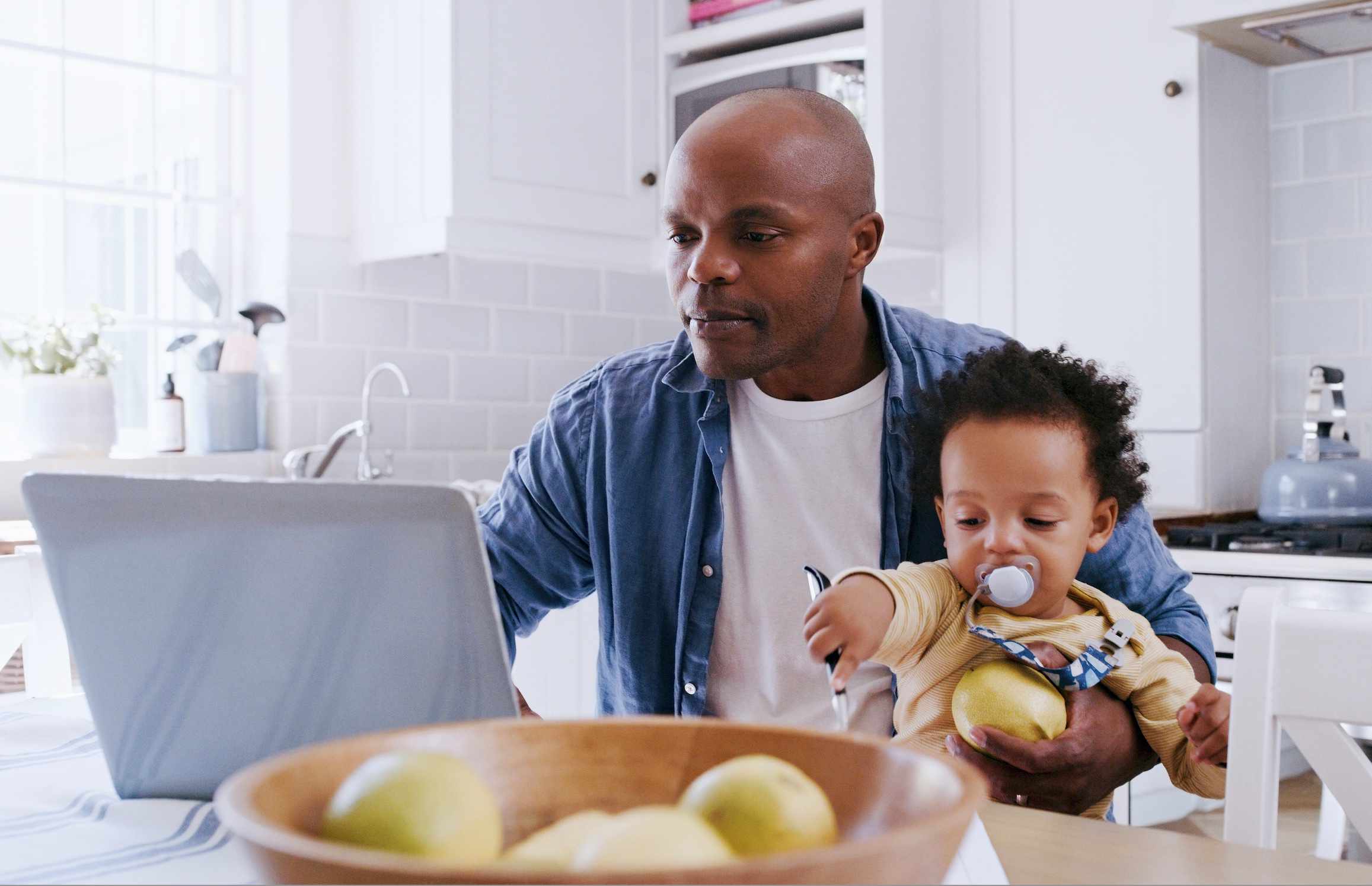 Black man in his 30s holding a toddler on his laptop while looking at his laptop at his kitchen table