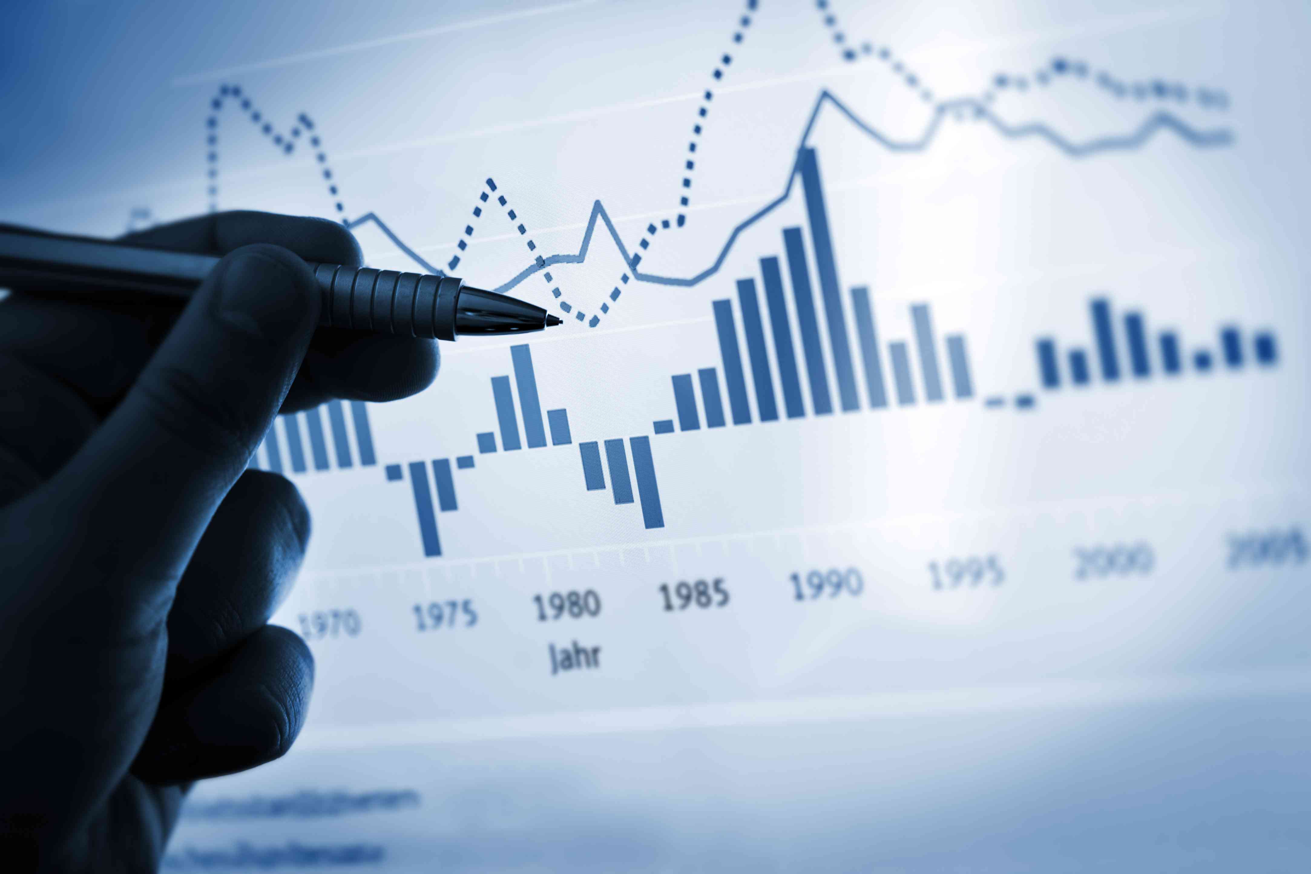 A hand with a pen pointing at a screen with financial charts.