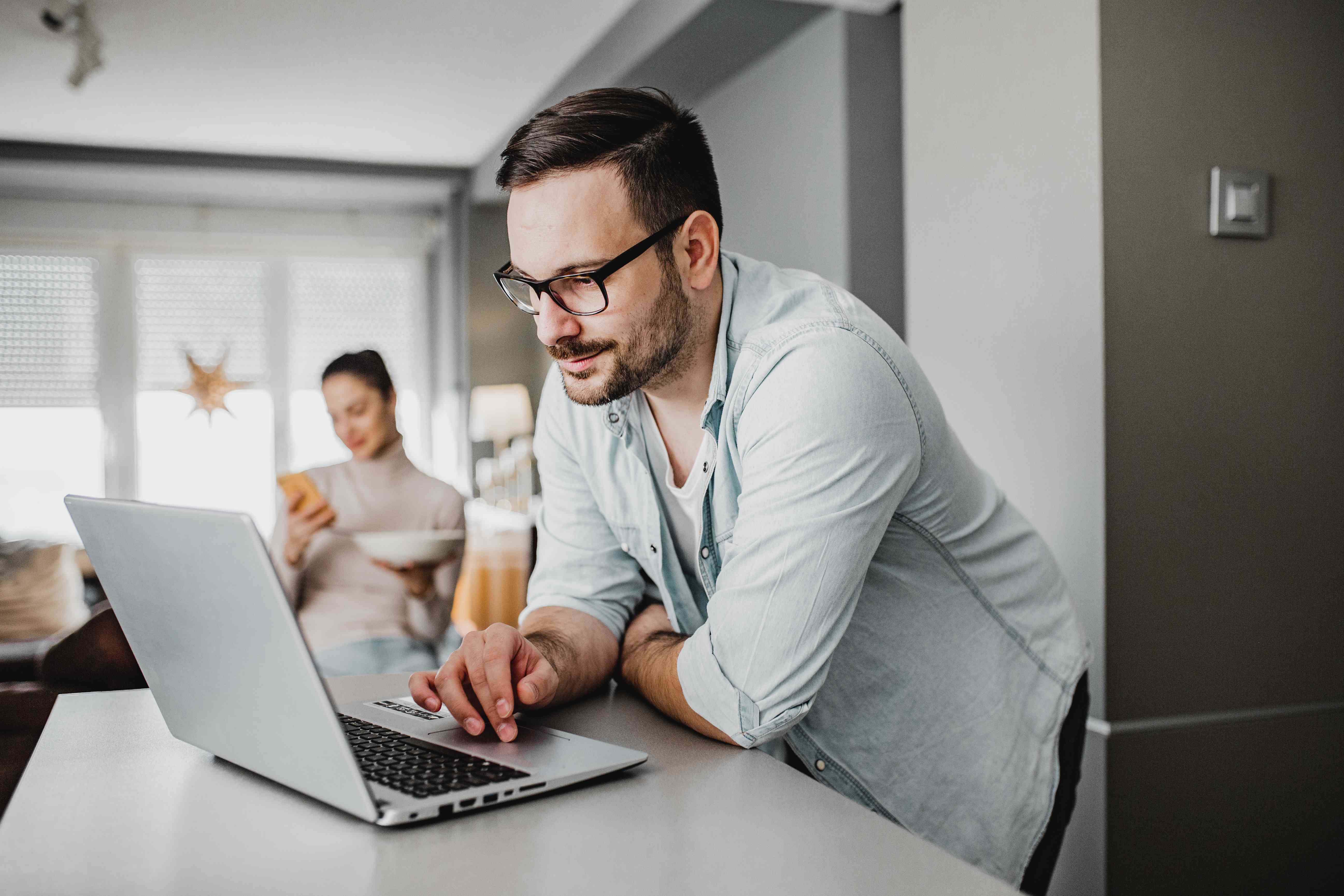 Man in his 30s at his kitchen countertop looking at his laptop, with his wife in the background