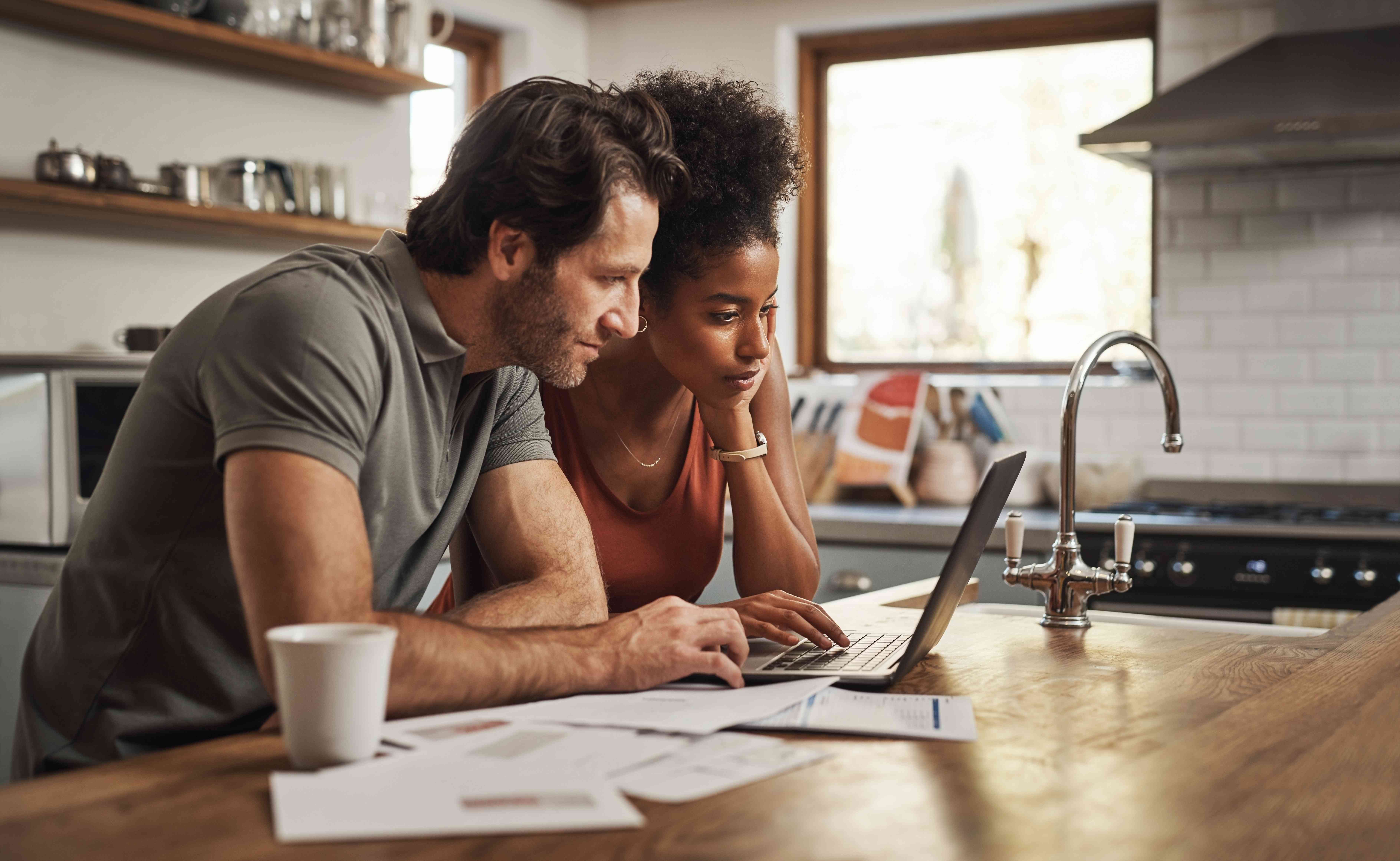 Young couple at their kitchen island looking together at a laptop with financial papers spread out