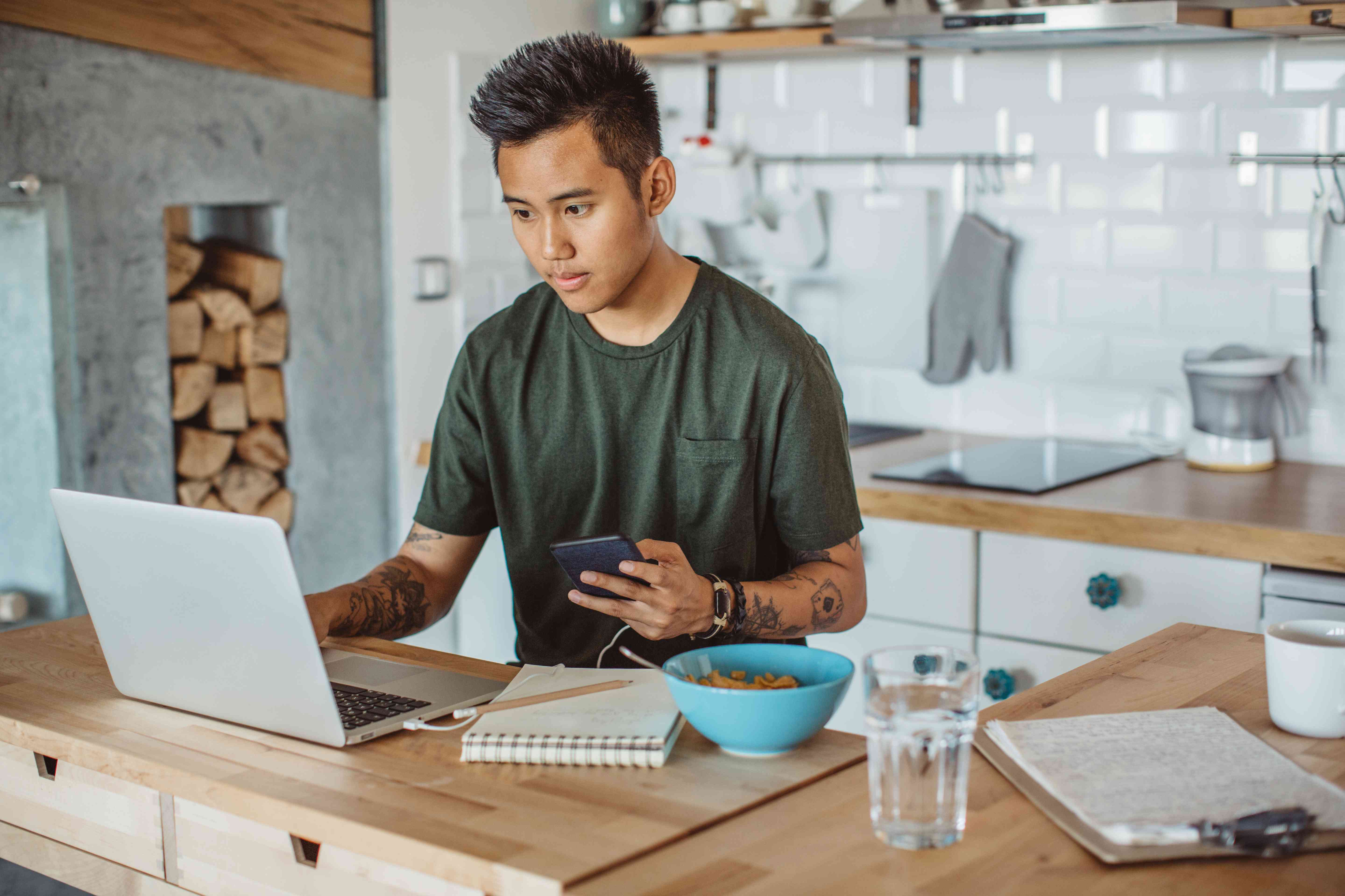 Young man sitting at his kitchen counter looking at his laptop and his phone, with a pencil and paper near him