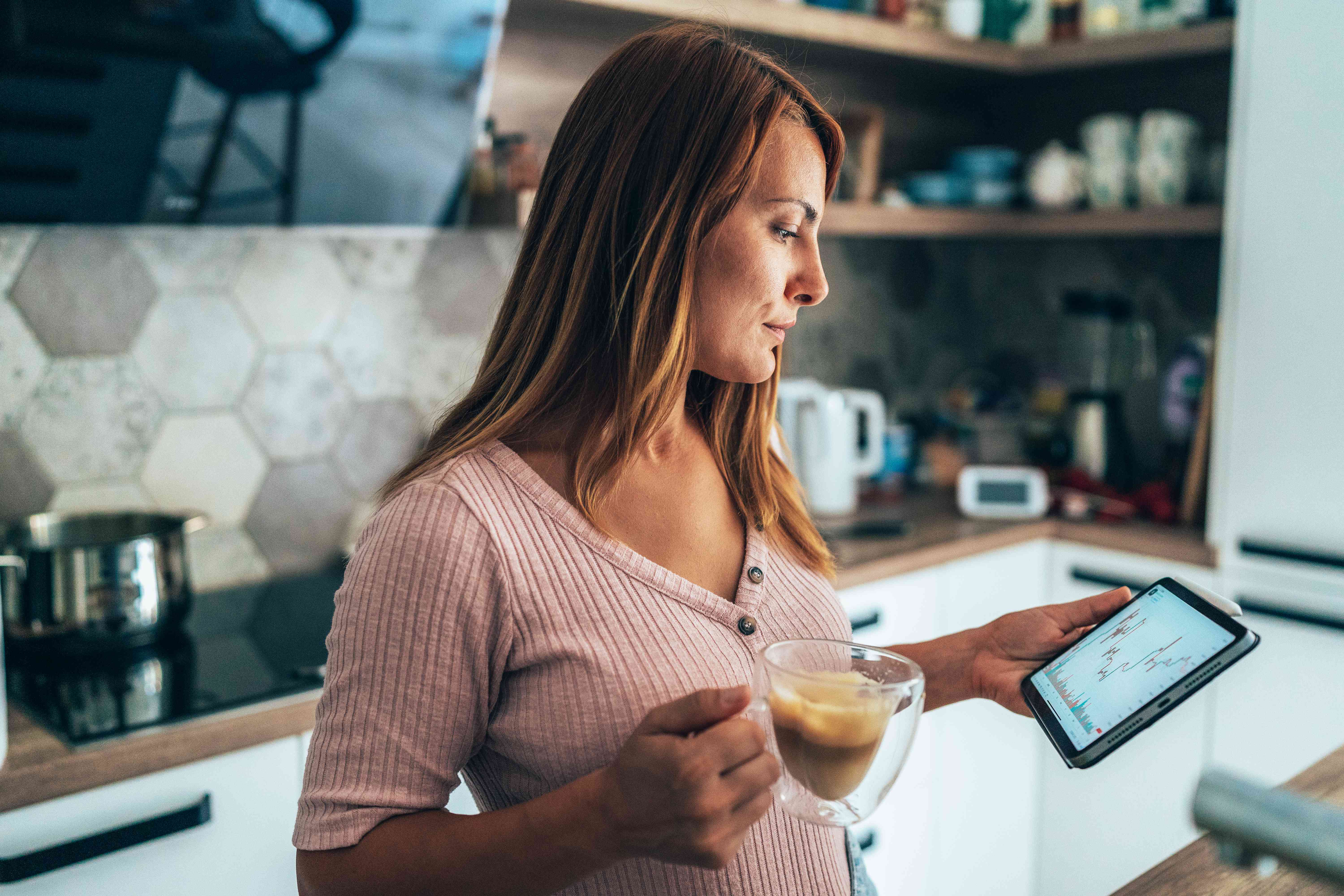 Woman looking at her trades on a handheld screen while holding a cup of coffee