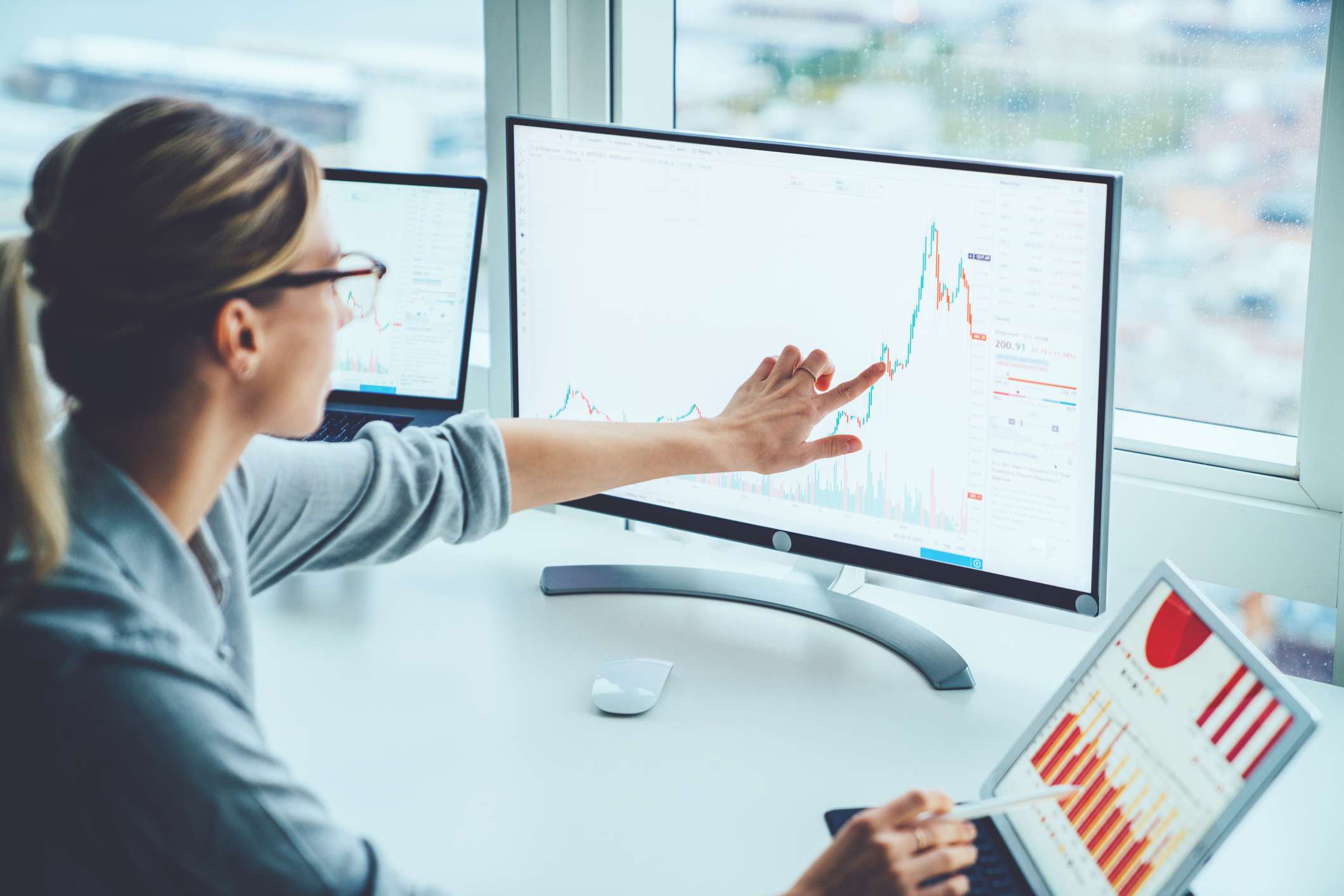 Businesswoman looking at touching a computer monitor showing a line graph with a laptop next to her showing bar graphs