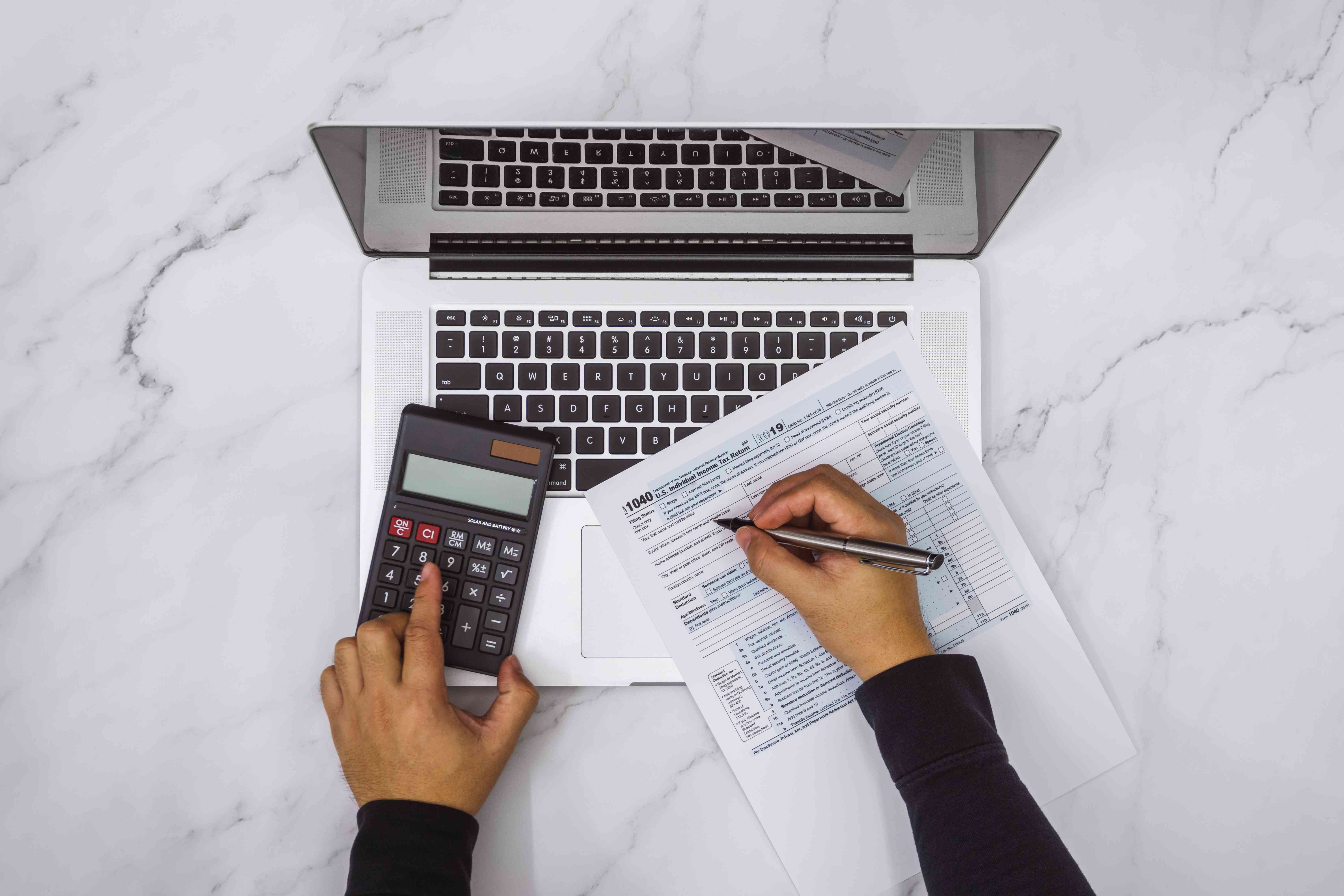 A person fills out a tax form and uses a calculator in front of a laptop.