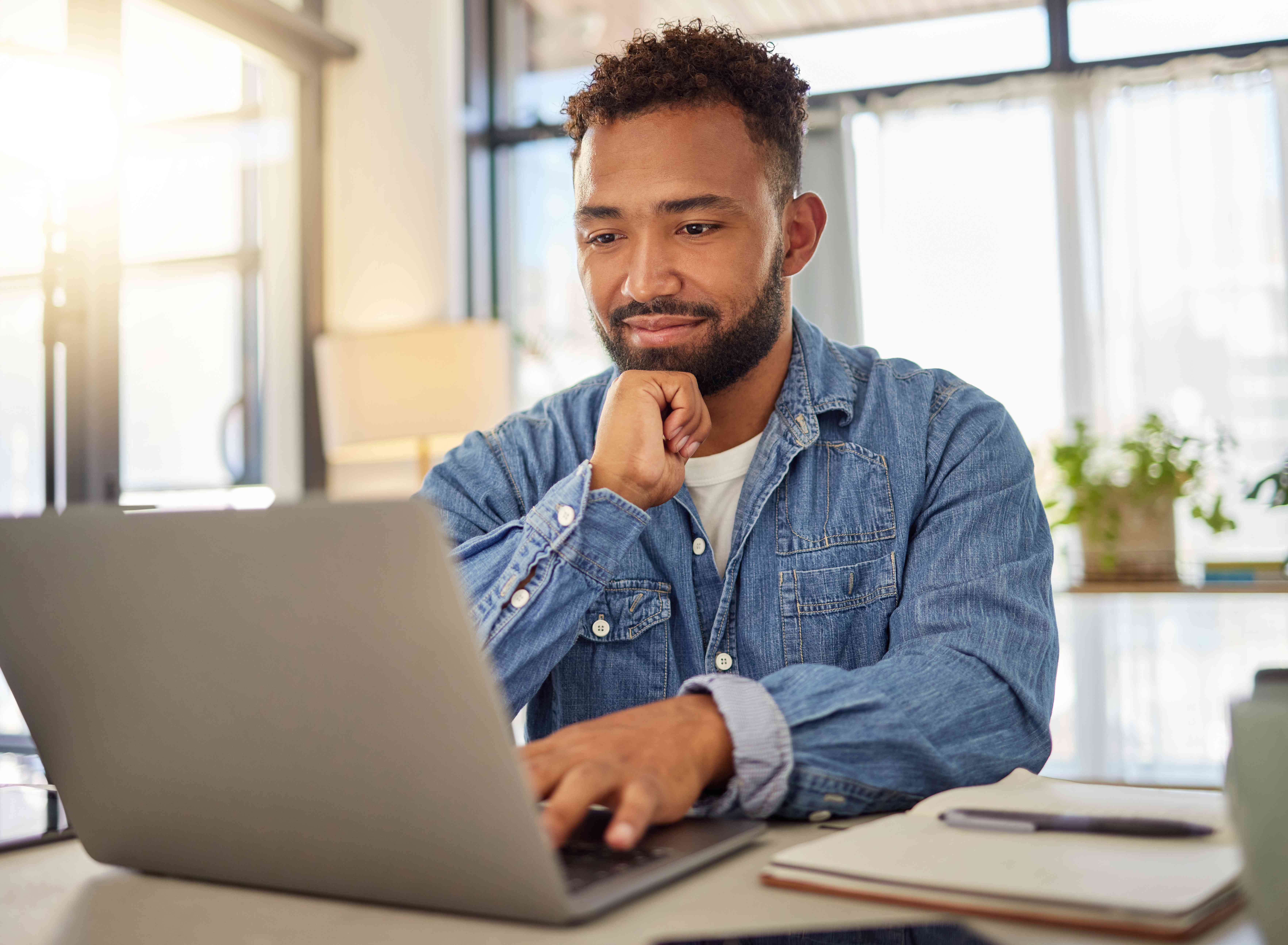 Young man at his kitchen table mildly smiling at his laptop with a notebook and pen nearby