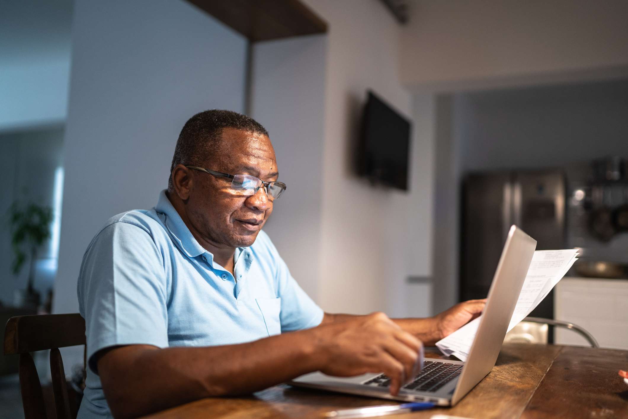 Older Black man at a kitchen table with a laptop holding paperwork