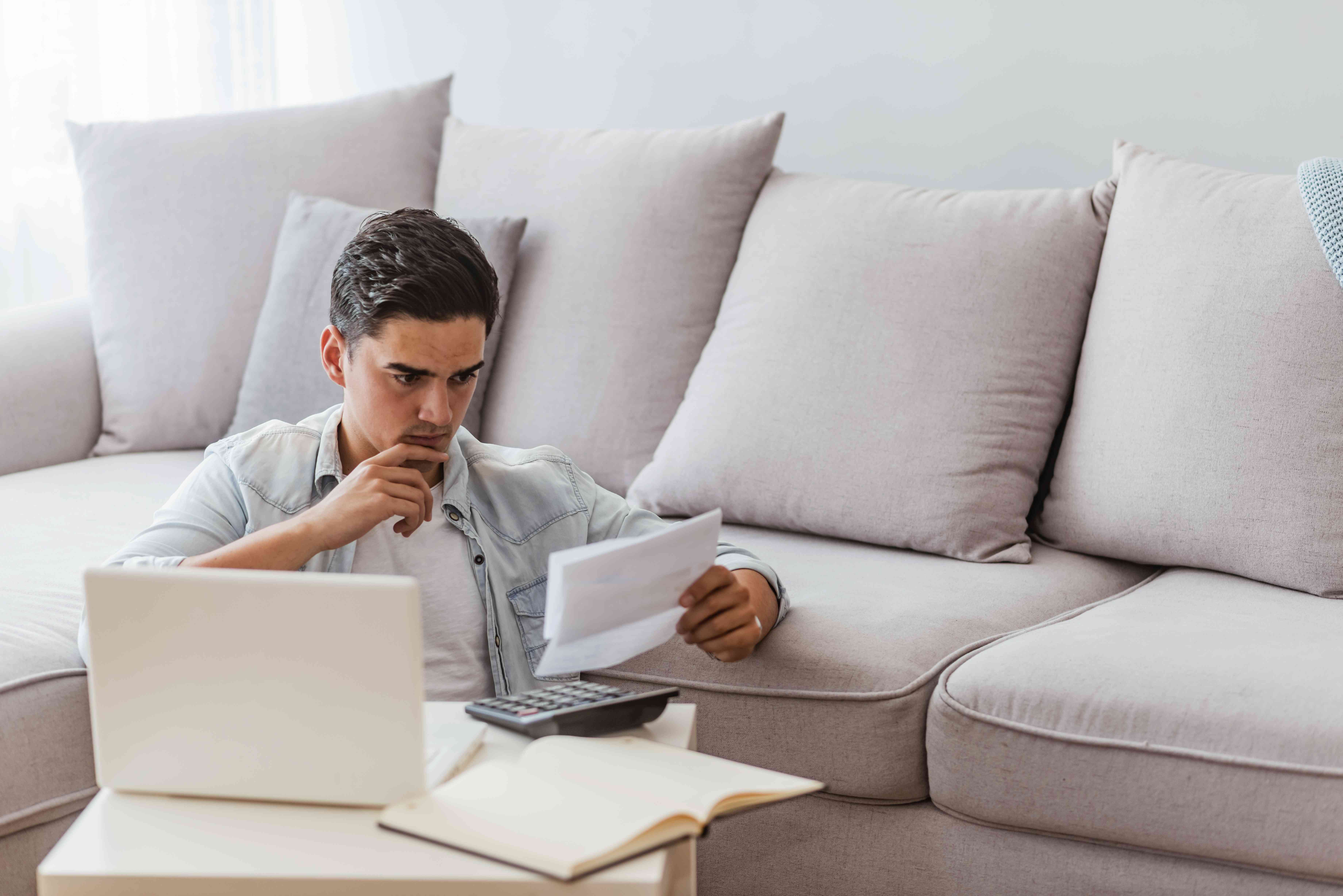 A thoughtful worker sits on the floor against a sofa and uses a calculator as they review their pay stub showing before and after tax wages.