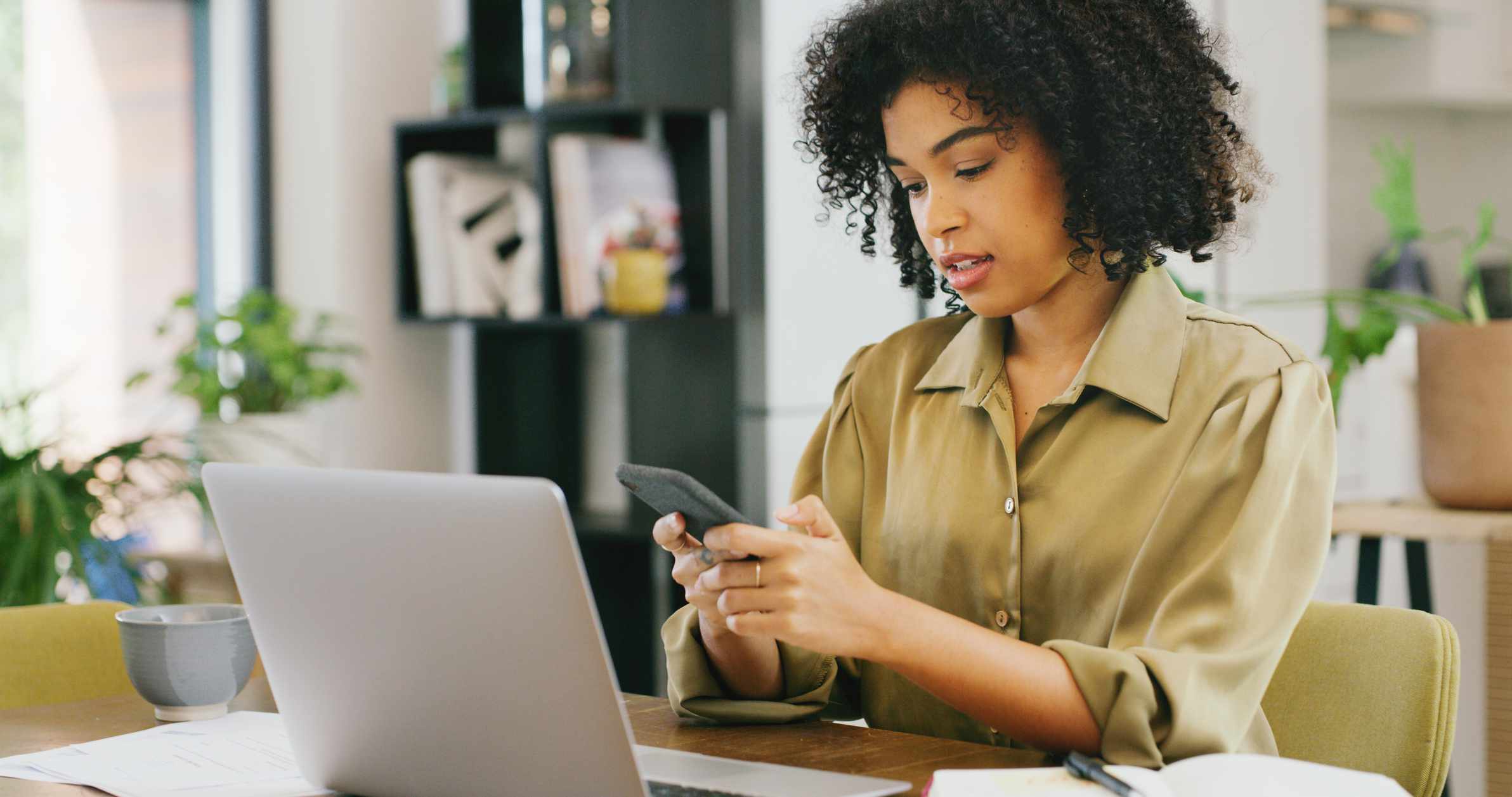 Woman using a smartphone and a laptop at a kitchen table.