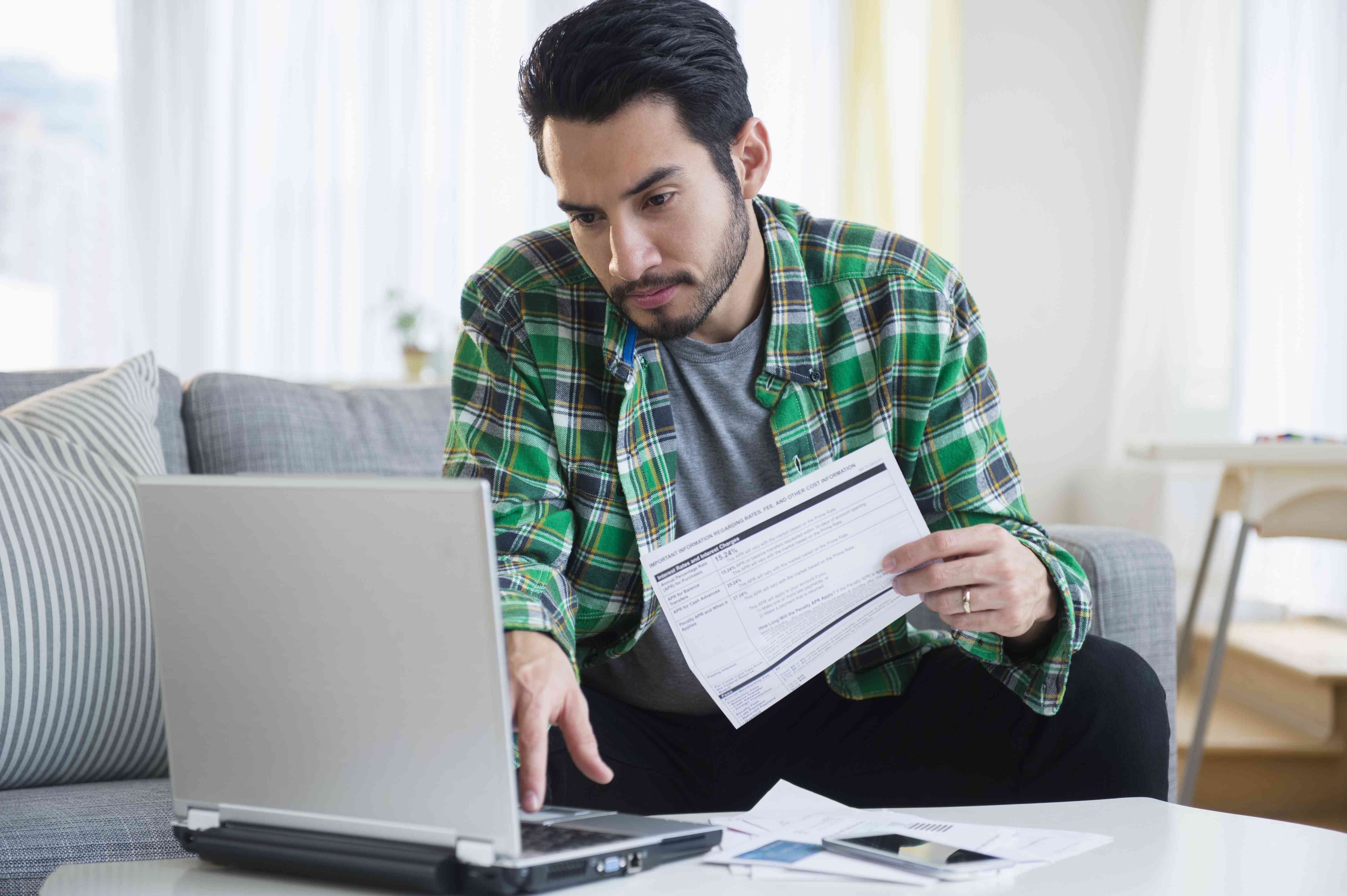 Man in his 30s at home on the couch, looking at financial documents and his laptop