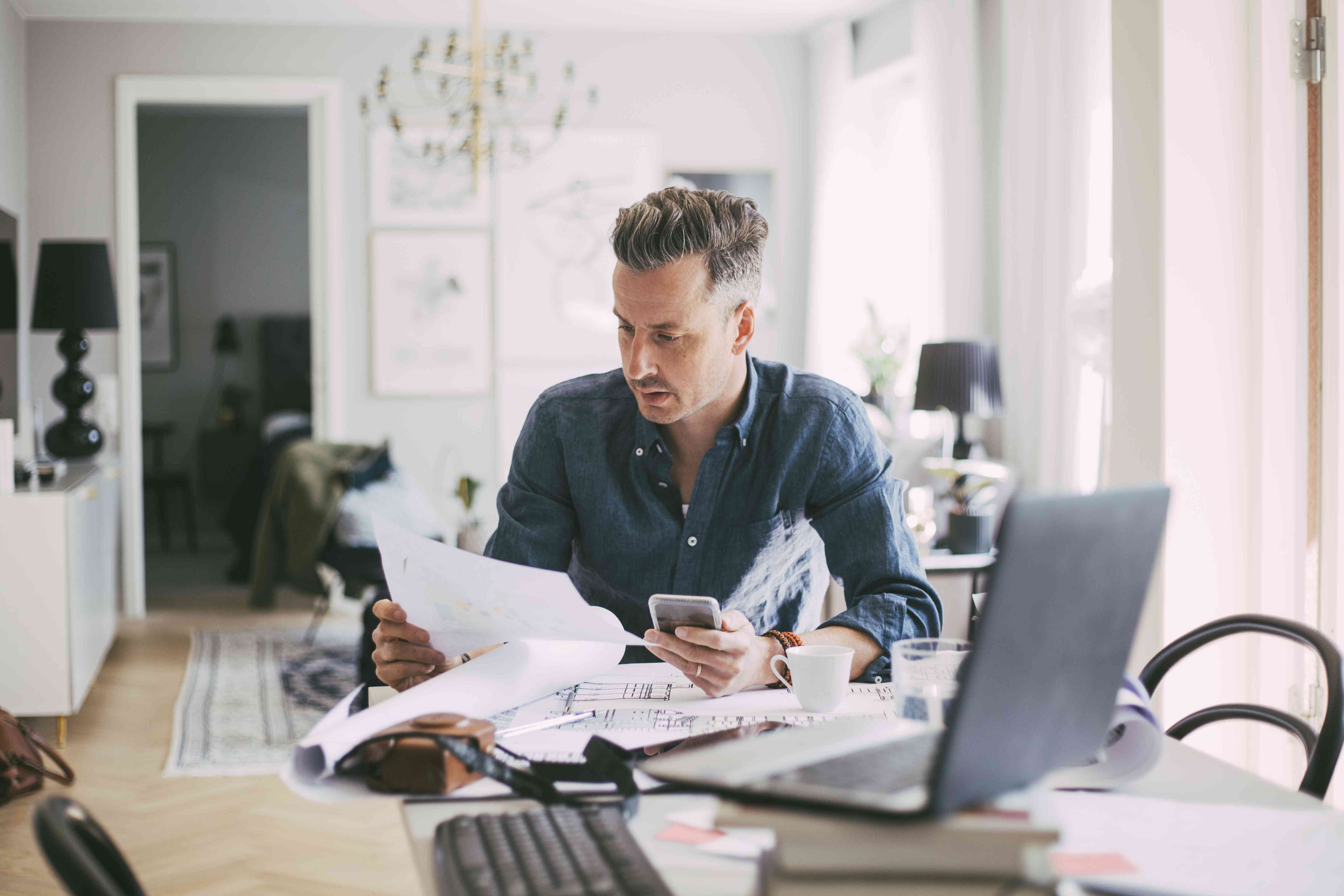 Man in his 40s at his dining room table looking at financial documents and a calculator, with an open laptop nearby