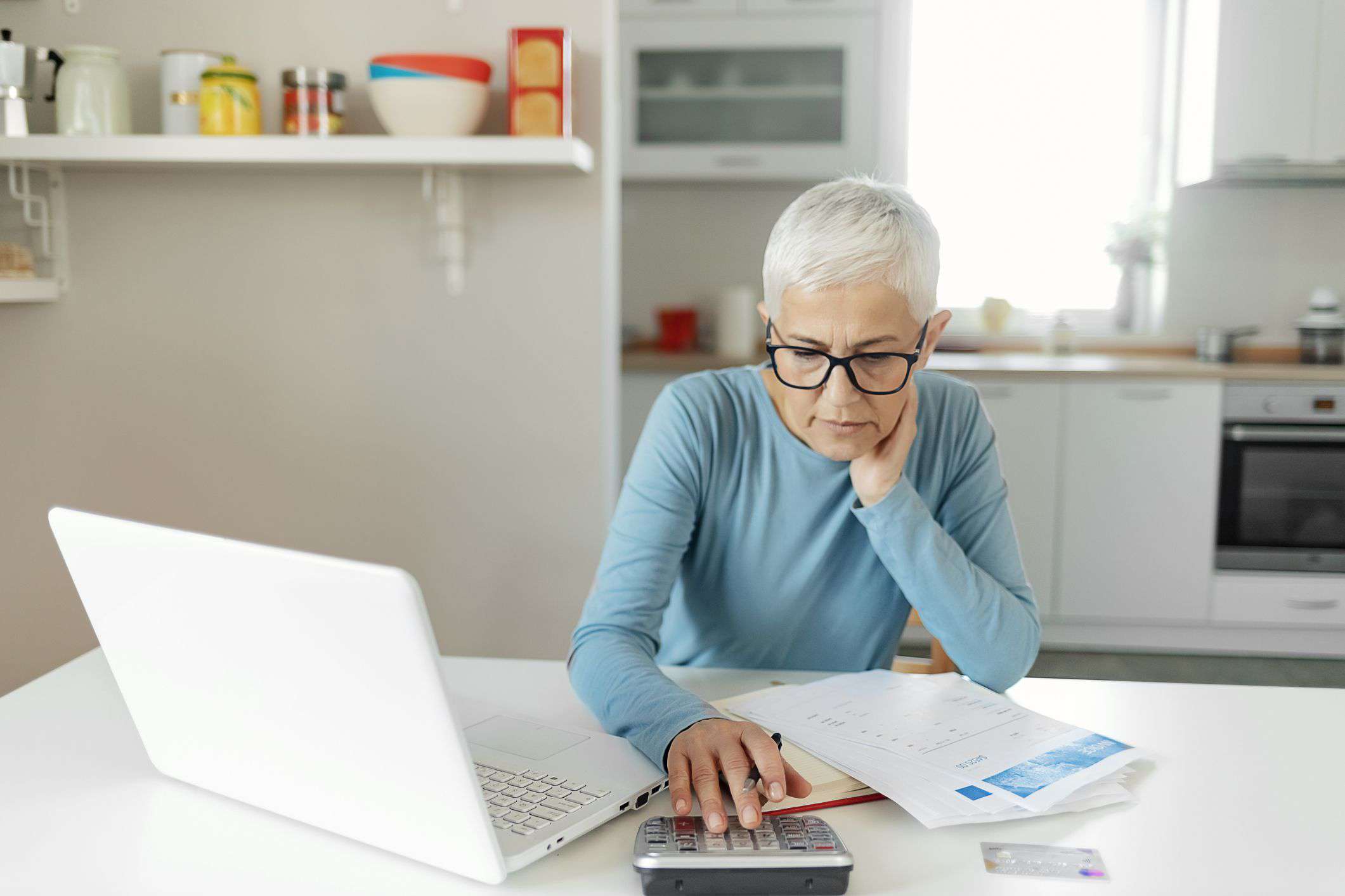 Woman with short gray hair in a blue top working with a calculator and laptop in her kitchen.