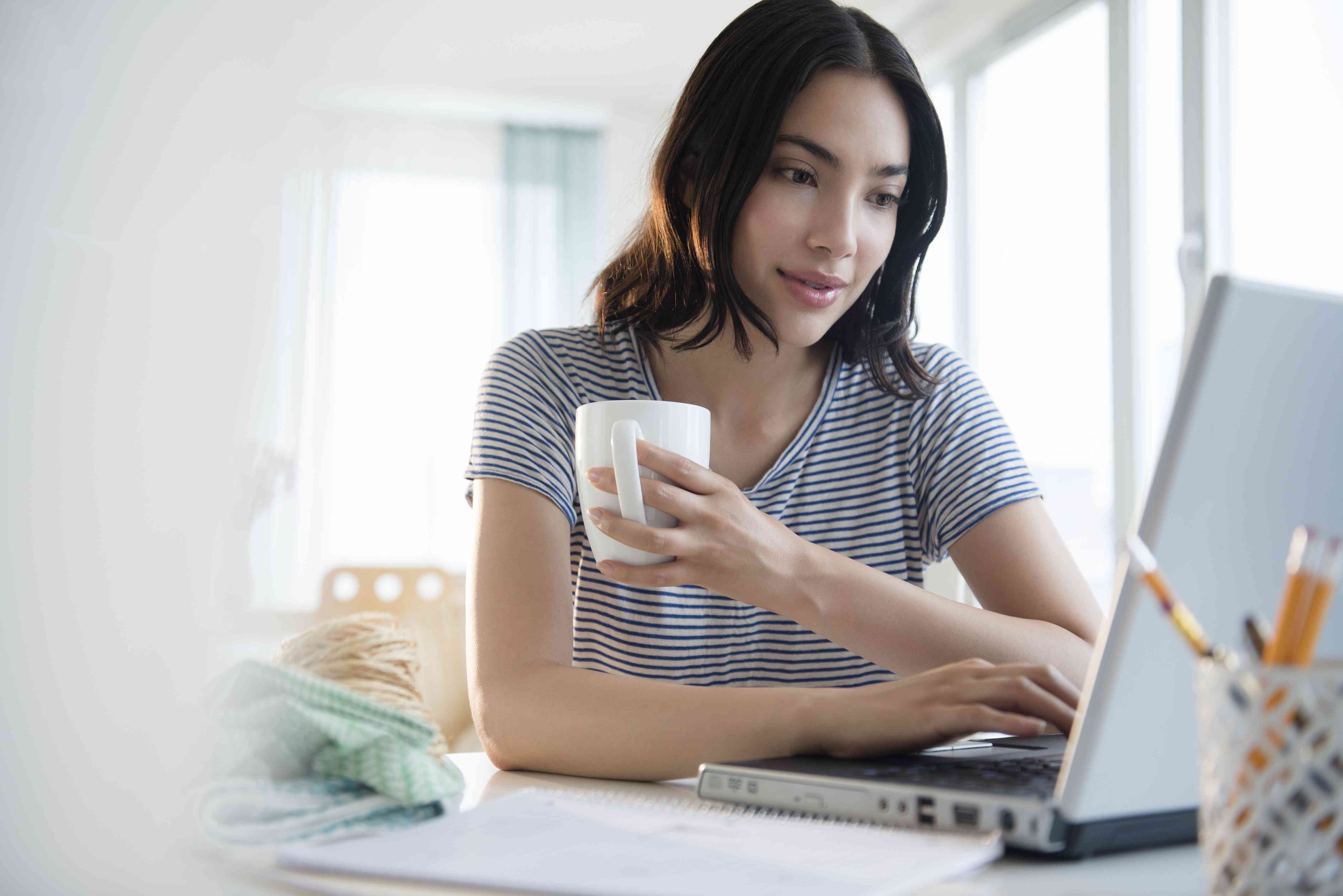 Young woman at home holding coffee cup while looking at her laptop