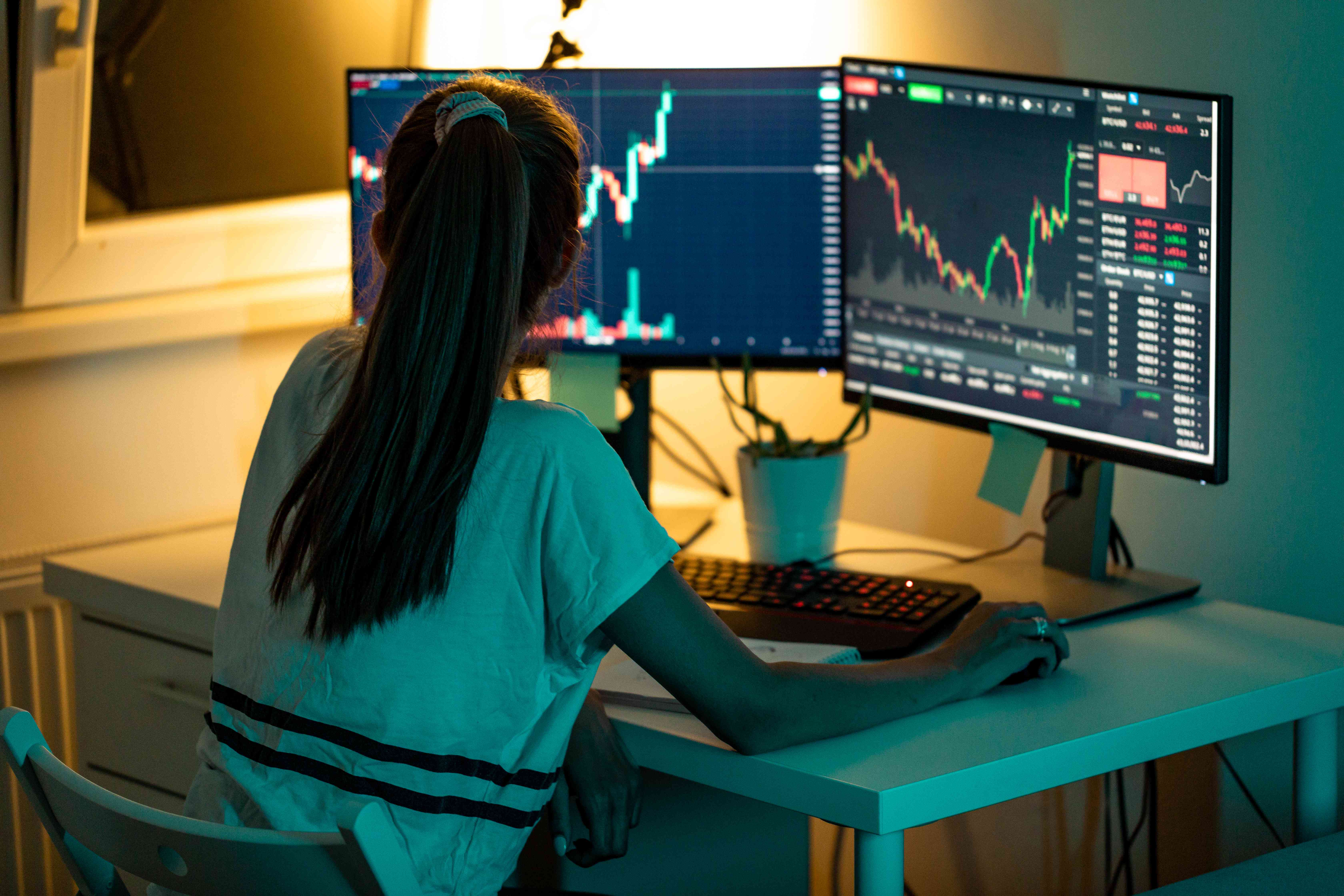 A woman sitting at a desk with her back to the camera looks at two computer screens displaying stock charts.