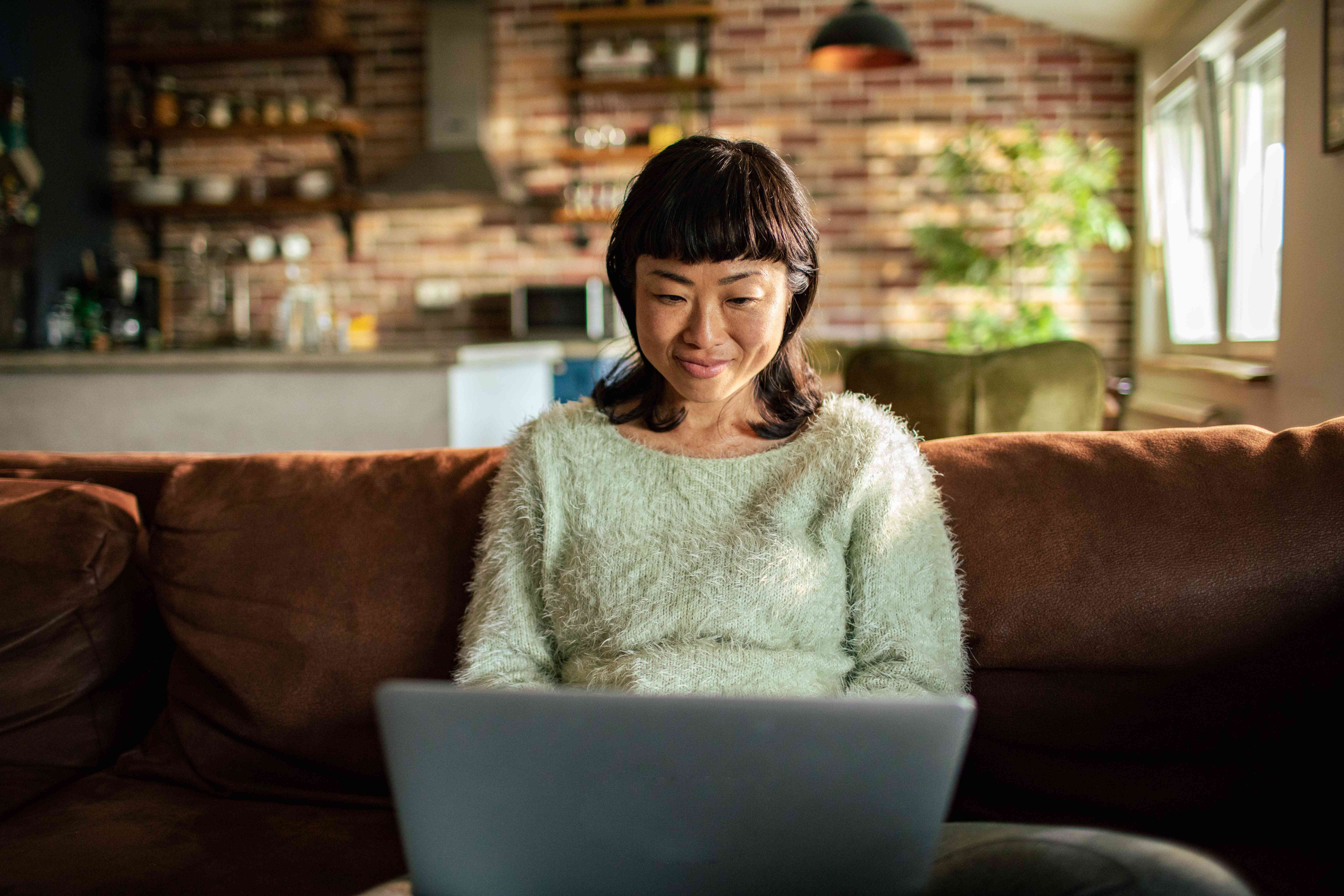 Young Asian woman sitting on her couch and smiling as she looks at her laptop