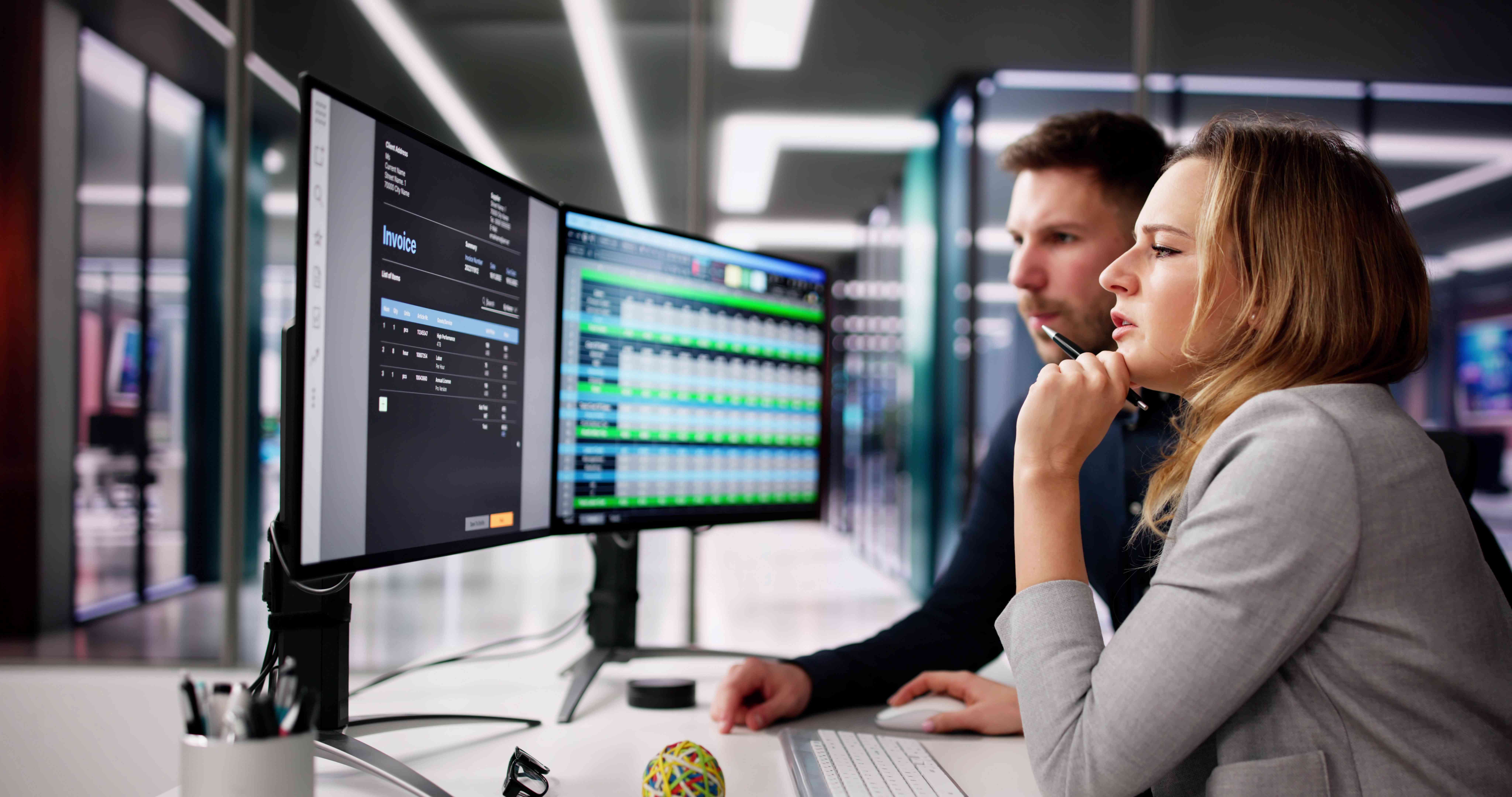 Two professionals look at two computer screens.