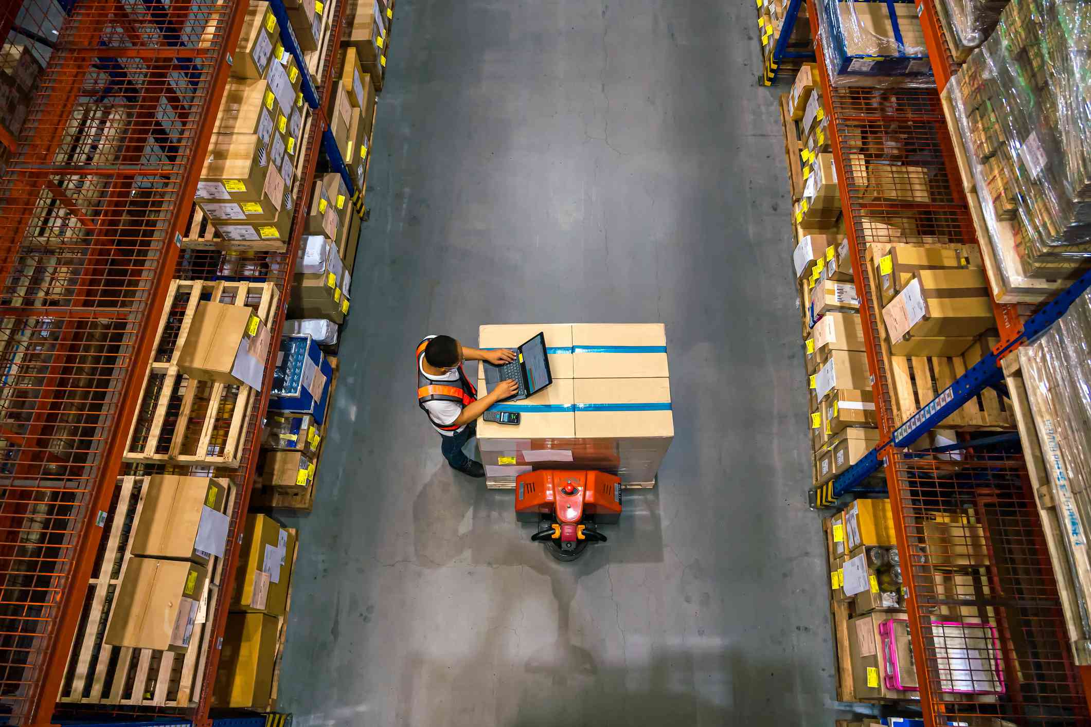 An aerial view of a warehouse worker typing on a laptop on top of boxes.