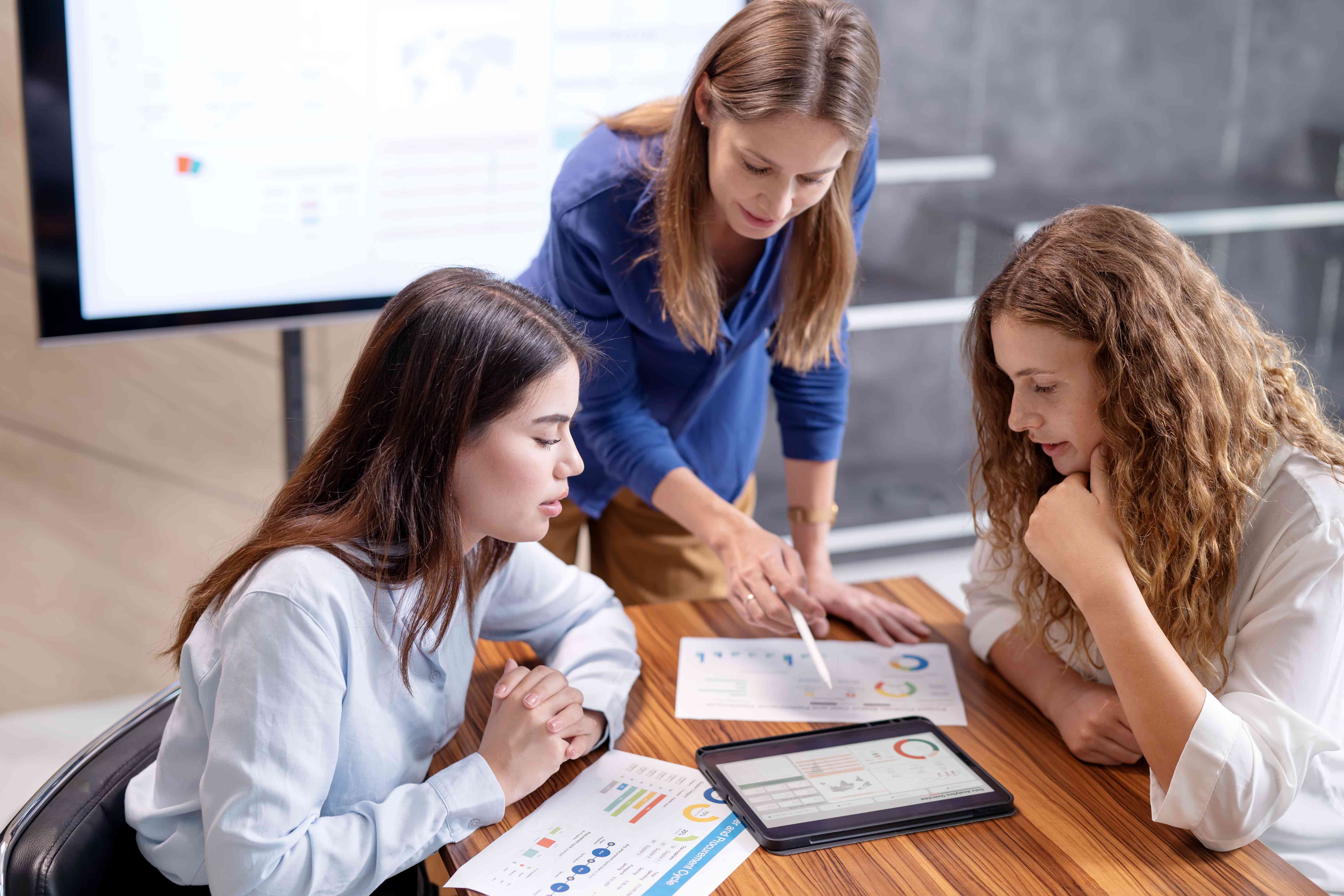 Three women in a conference room. One standing over them discussing a document.