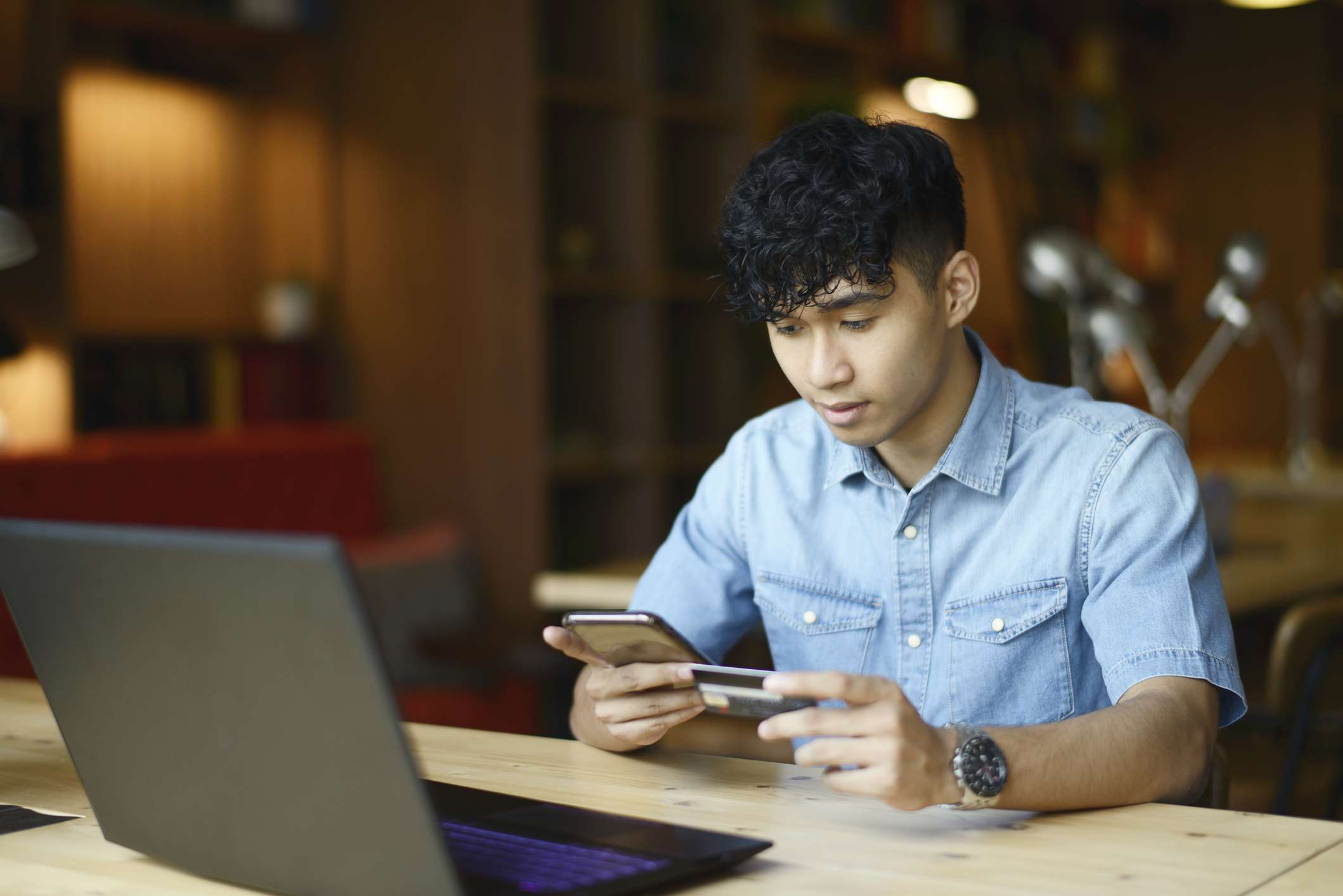 a young person sitting at a desk in front of an open laptop, looiking intentently at a phone held in one hand and a credit card in another