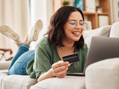 A happy woman lies on a couch while holding a credit card and looking at a laptop. 